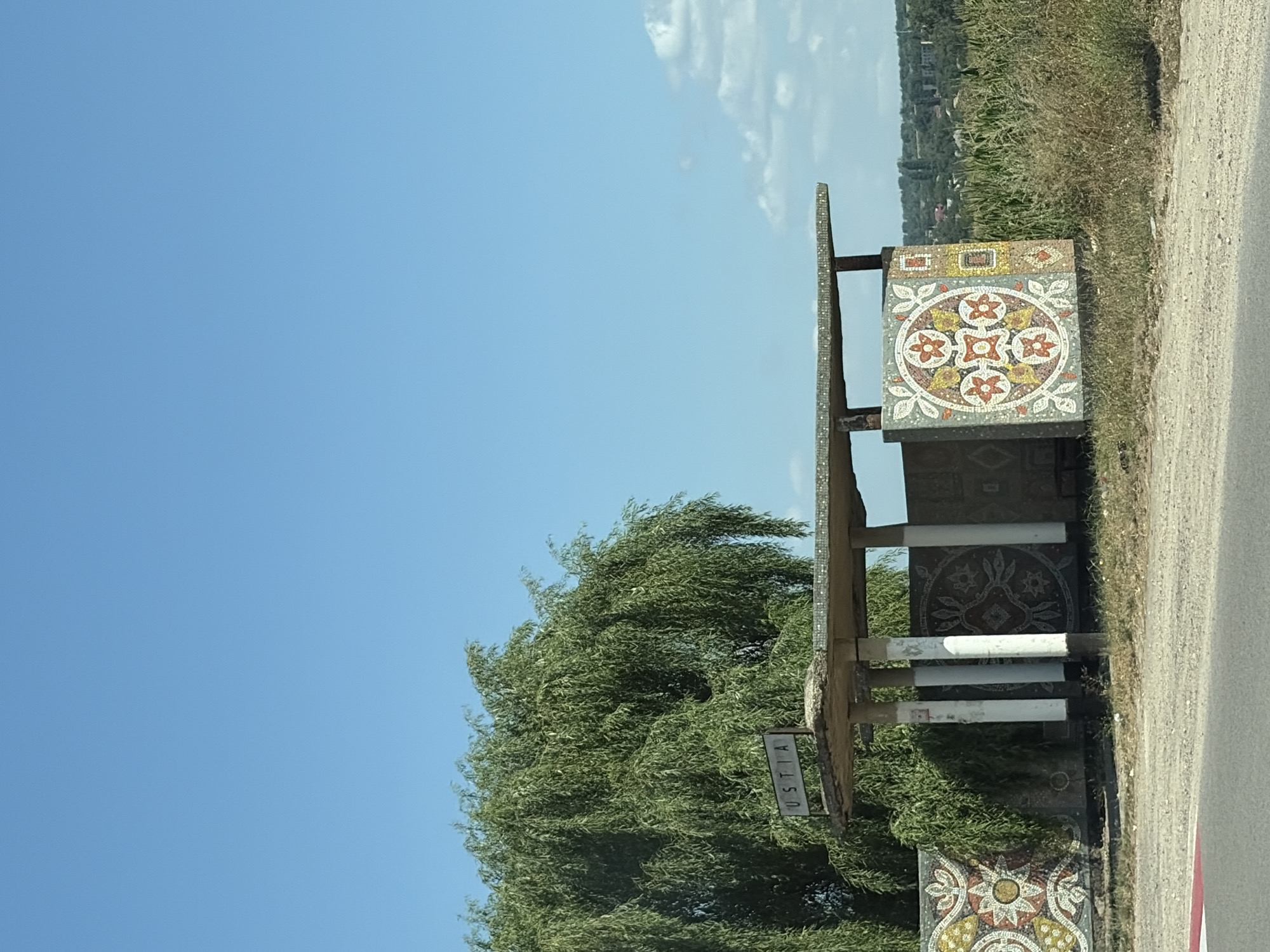 The image depicts a bus stop located in the city of Ustia, Moldova. The bus stop features a simple concrete roof supported by columns, adorned with decorative mosaics. A sign reading "USTIA" is visible above the shelter. The surrounding area is characterized by a large, leafy tree, a mosaic wall, and vegetation, including corn stalks in the background. The sky is clear blue with a few scattered clouds. The ground appears to be a dirt road with a paved section in the foreground.