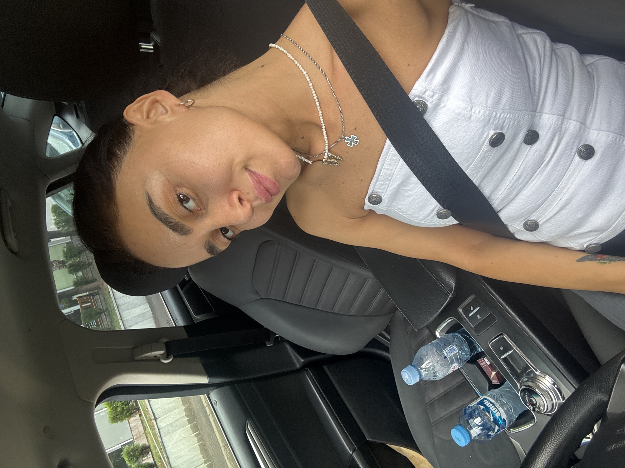 The image shows a woman sitting in the driver's seat of a car. She is wearing a white strapless top and a seatbelt. There are two bottles of water and a pack of cigarettes on the center console. The car appears to be parked on a street in Tbilisi, Georgia. The background shows buildings and trees.