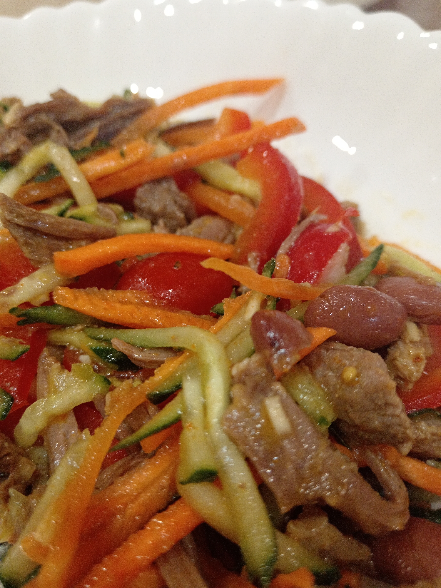 A close-up shot focuses on a vibrant salad served in a white, scalloped bowl. The salad is a medley of shredded vegetables and thinly sliced meat, possibly beef, intermingled with kidney beans and cherry tomatoes. Prominent ingredients include julienned carrots, zucchini, and red bell peppers, all tossed in a light dressing that gives them a glistening appearance. The dish is presented in a well-lit indoor setting, suggesting it's a prepared meal rather than a street food offering. The context of Uchkeken, Russia, is not evident from the visual information provided in the image itself.