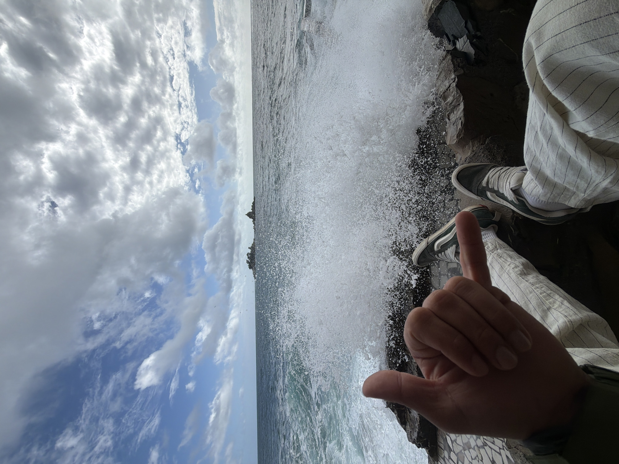 A person is sitting on a rocky cliff at Petrovac centar, Montenegro, with waves crashing against the rocks. The person is wearing striped pants and sneakers. They are holding up a "hang loose" hand gesture. The sky is partly cloudy.