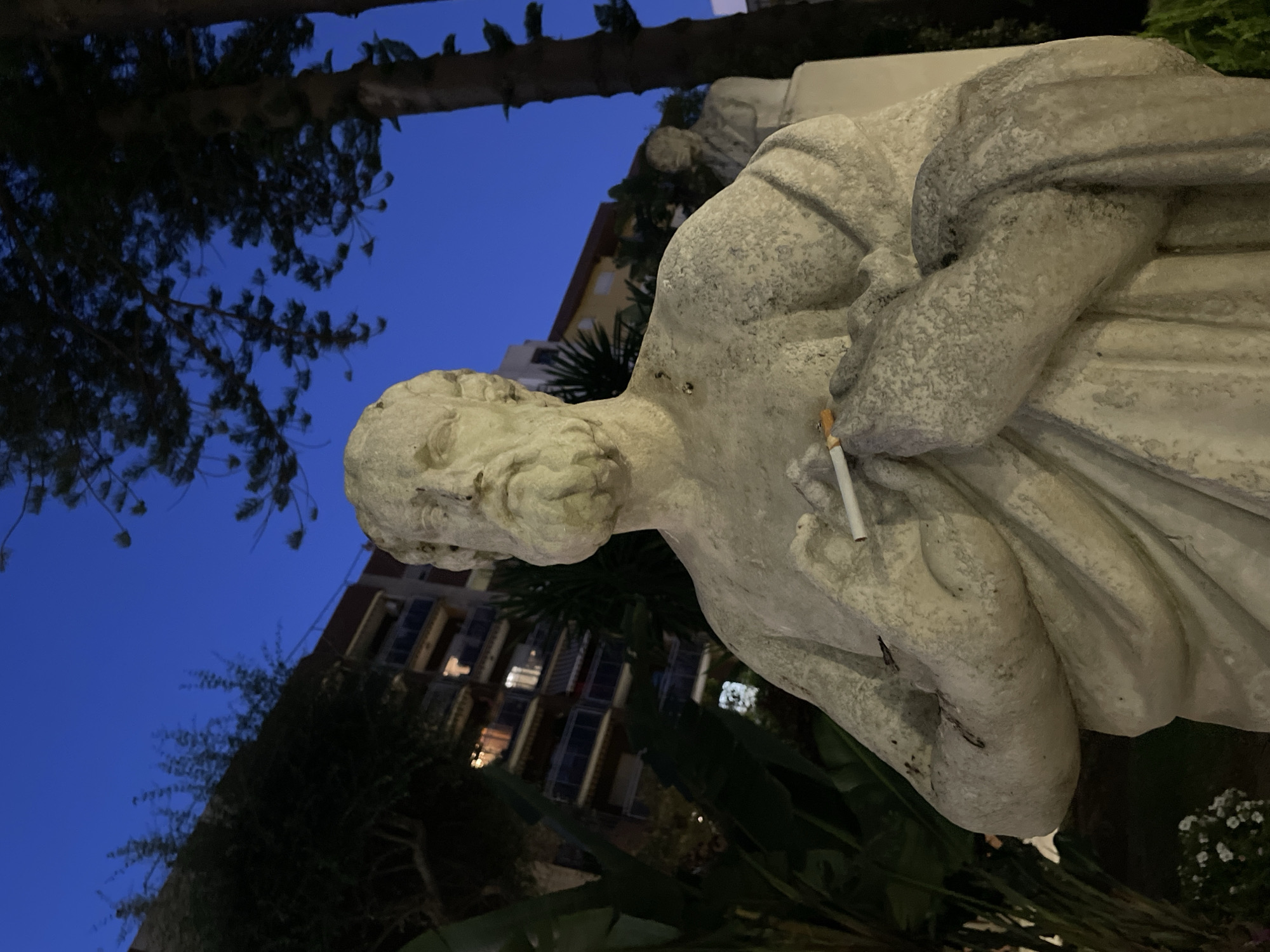 A close-up, low-angle shot shows a weathered, stone statue of an elderly man with a beard, wearing a toga. The statue is holding a lit cigarette between its right thumb and forefinger. The statue is positioned outdoors at dusk, with a deep blue sky visible in the background. Behind the statue, a multi-story building with illuminated windows can be seen, along with dark green foliage and the trunk of a pine tree. Another, less distinct statue is visible in the background to the right. This scene is located in the city of La Línea de la Concepción, Spain.