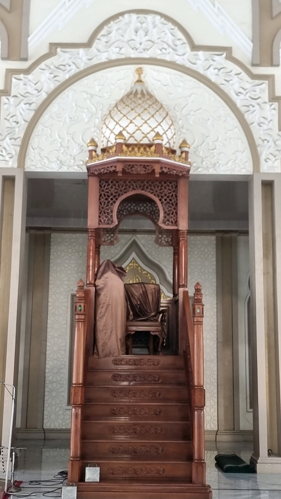 The image shows a wooden minbar, or pulpit, inside a mosque. The minbar is ornately carved and features a dome on top, decorated with gold accents. A chair is placed on the pulpit, covered with a brown cloth. The mosque interior is decorated with patterned wallpaper and intricate architectural details. A prayer mat is visible on the floor to the right of the minbar. The image was taken in the city of Lhokseumawe, Indonesia.