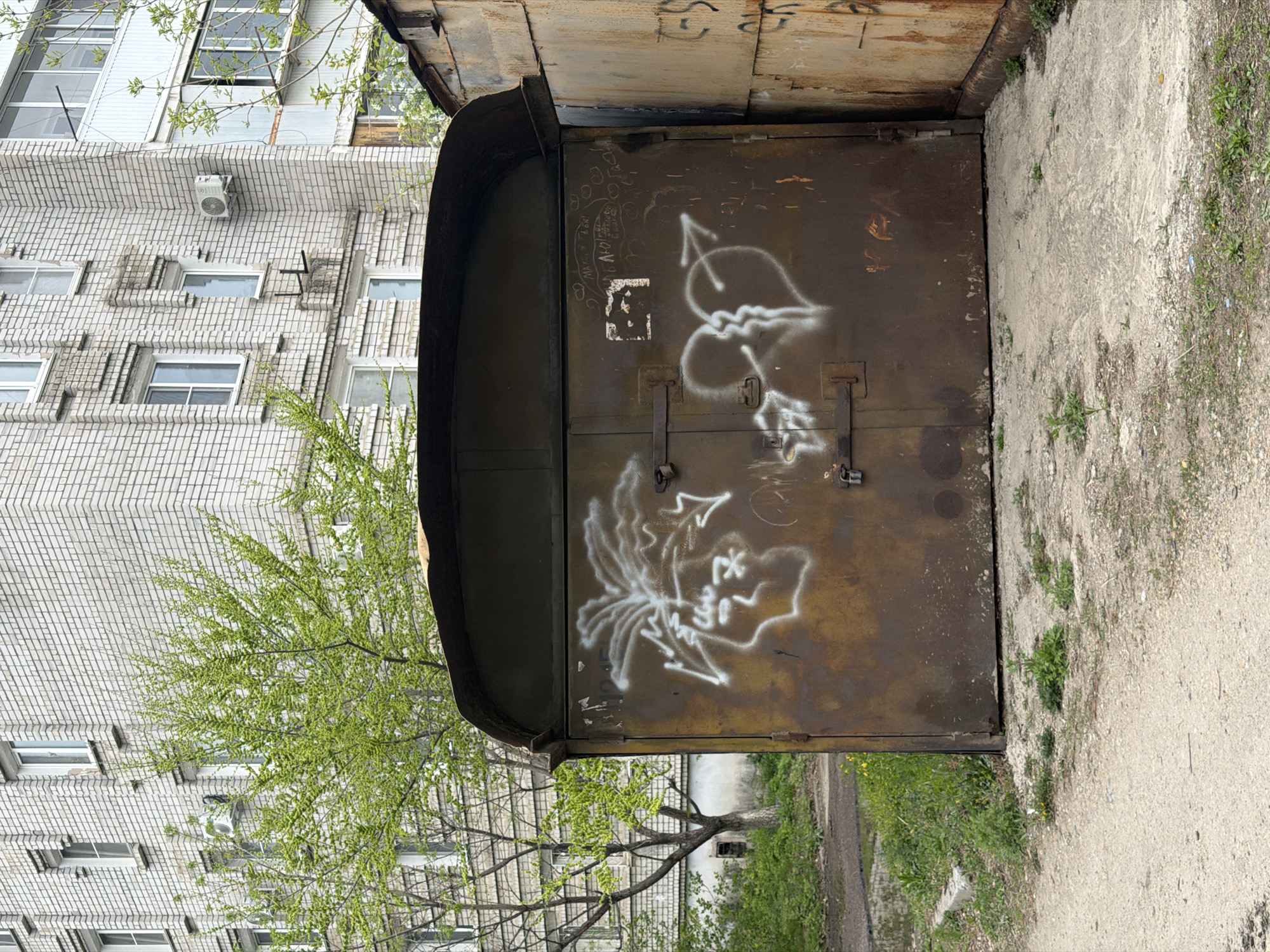 The image shows a corroded, brown metal garage door with white graffiti on it. The graffiti includes a heart shape with an arrow and abstract designs. The door is secured with metal latches and padlocks. Above the door, there is a curved metal overhang. To the left, a tree with fresh green leaves is partially visible, in front of a light-colored brick building with multiple windows. To the right, there is a concrete walkway with weeds and grass growing along the edges. The location is possibly in Birobidzhan, Russia, based on the prompt's instruction.