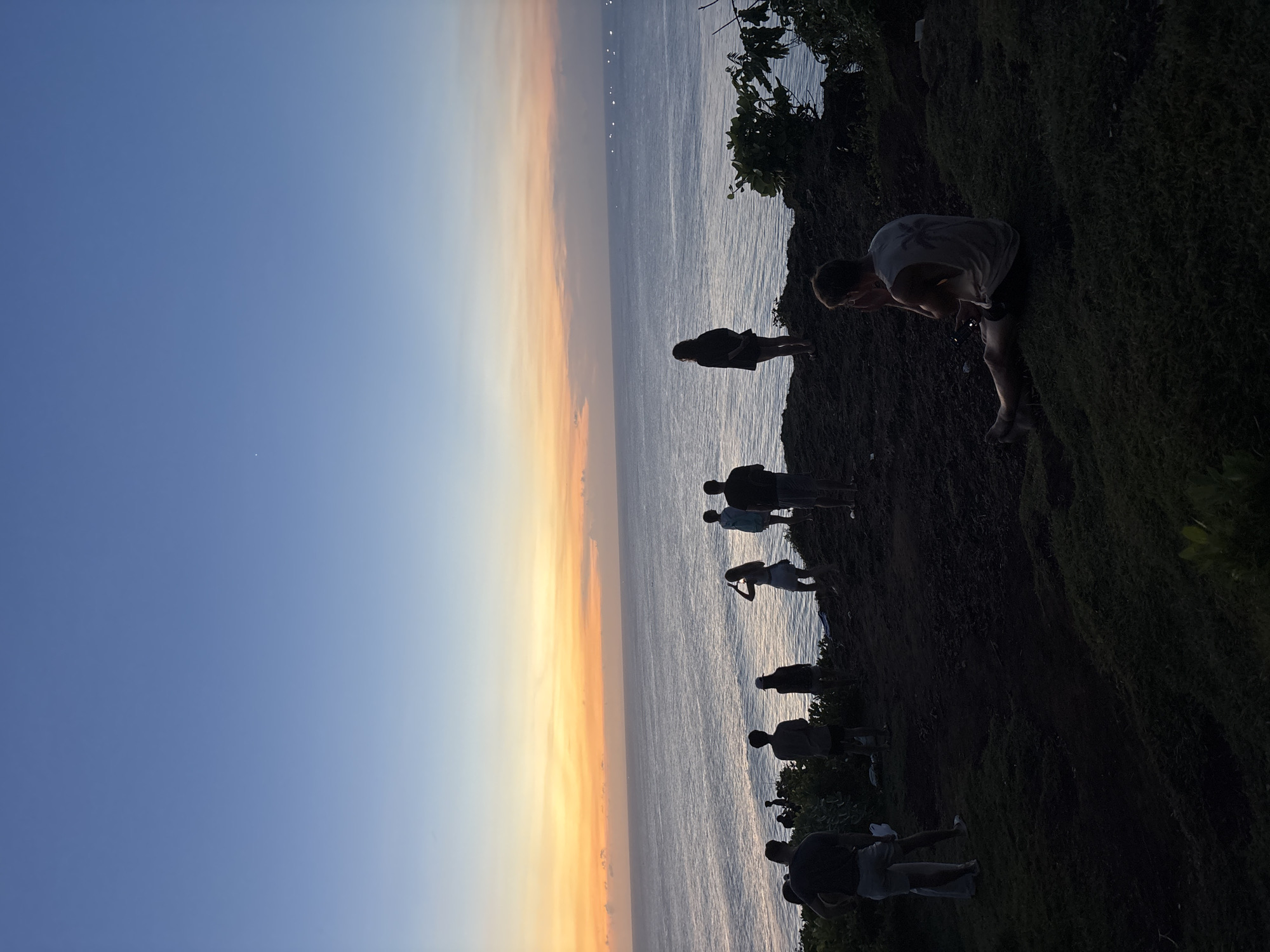 The image shows a group of people standing on a cliff overlooking the ocean at sunset in Kuta Selatan, Indonesia. There are approximately ten people visible, mostly silhouetted against the light. One person is leaning over the edge of the cliff, possibly taking a photograph. The sky is a gradient of blue to orange, reflecting on the water's surface. Vegetation is visible on the cliff face. The ground is covered in grass and appears uneven.