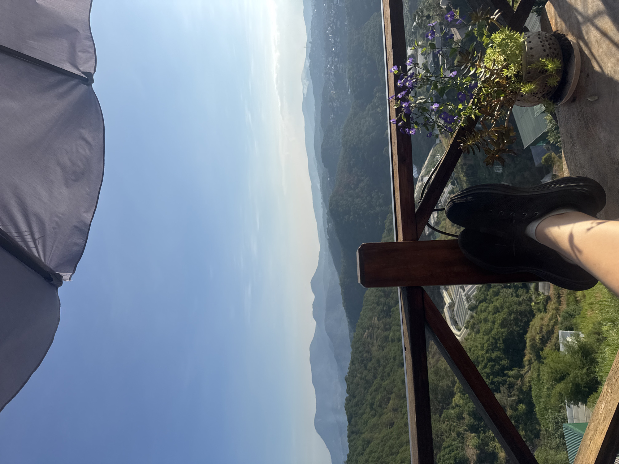 The image depicts a view from a balcony overlooking a mountain range, likely in Da Lat, Vietnam. The scene includes a clear blue sky, a landscape with rolling hills, and a city skyline in the distance. A portion of a dark-colored umbrella is visible on the left side. On the balcony, a wooden railing is present, with a potted plant featuring purple flowers on the side. A pair of black shoes is placed on the railing, and a pale leg wearing a white sock is also seen. The environment suggests a bright, sunny day. The perspective is from a person sitting on the balcony, enjoying the view. The image does not depict actions or interactions.
