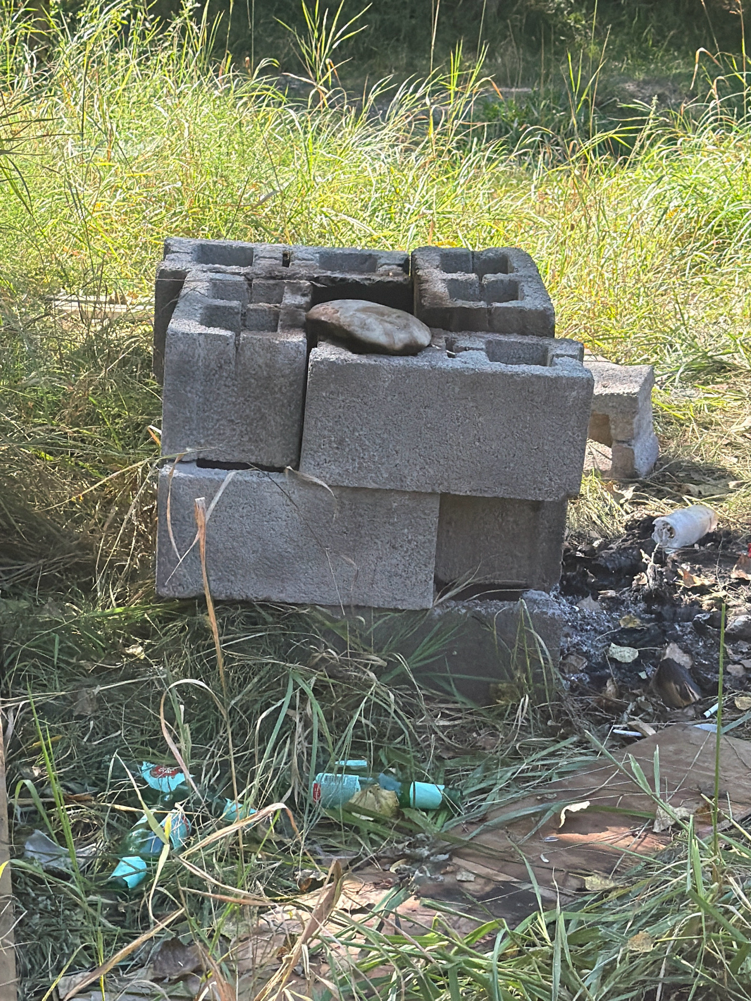 The image shows an outdoor scene with overgrown green grass and a crude fire pit constructed from cinder blocks. The fire pit is stacked unevenly, with a flat stone resting on top. There are signs of past fires, including ash and charred debris on the ground around the pit. Scattered in the grass are several empty plastic bottles, some appearing to be green or blue. The overall impression is one of neglect and disarray. This scene is located in the city of Yessentuki, Russia.