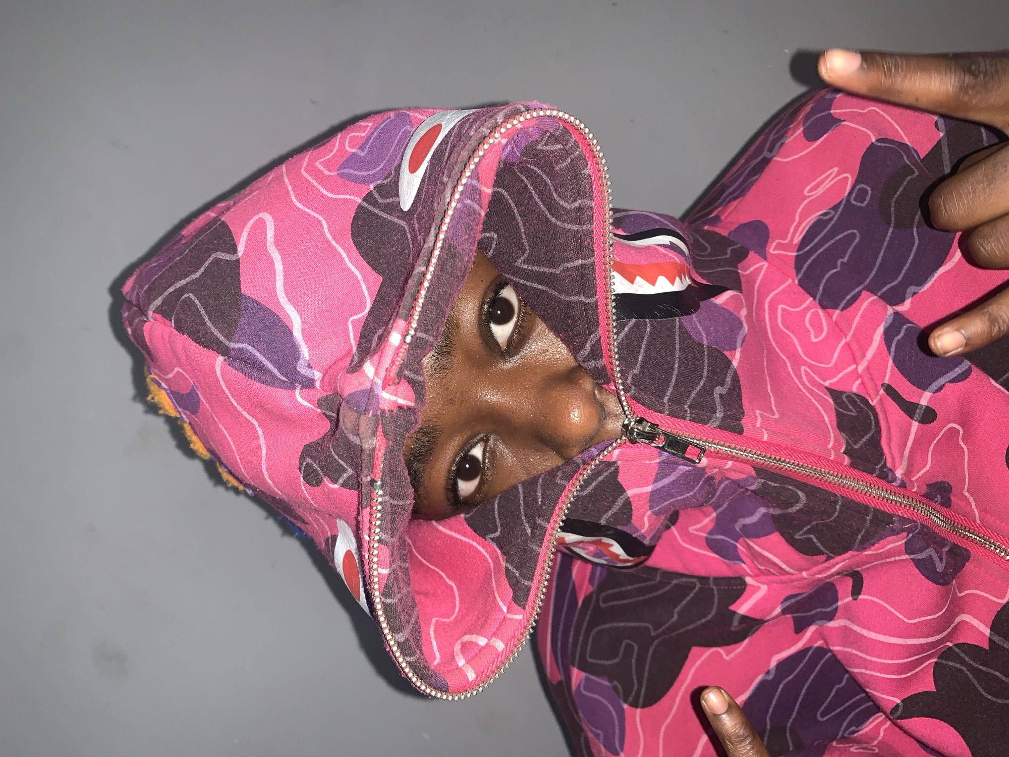 A person with dark skin is wearing a pink, purple, and brown camouflage hooded sweatshirt. The hood is up and partially zipped, framing their face, and only their eyes and part of their nose are visible. The person's fingers are visible on the right side of the image. The background appears to be a plain, gray surface. There is no discernible context indicating a specific location. Therefore, it's not possible to determine if this image was taken in Rafin Par, Nigeria.