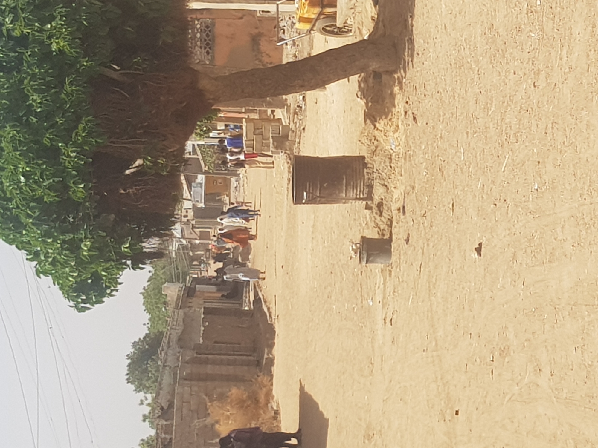 The image depicts a street scene in Dutsinma B, Nigeria. The unpaved road, dominates the frame, with a large tree on the left casting a shadow across it. Buildings line the left side of the road, some in disrepair. Several people walk along the road, their attire suggesting everyday activities. A metal barrel sits on the right side of the road. Overhead wires are visible against the sky in the upper left. The scene is bathed in bright sunlight.