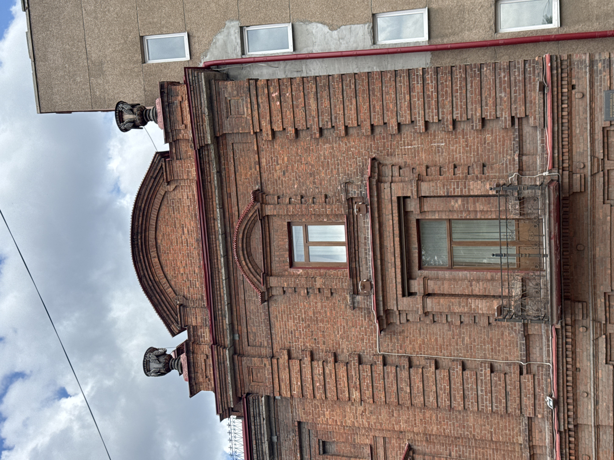 The image shows an exterior view of a brick building with a distinctive architectural style, likely from the early 20th century. The building is constructed with red bricks and features decorative brickwork, including projecting bricks arranged in patterns and a rounded architectural element above a window. There are two visible windows on the upper part of the building, one with white trim and the other with a darker frame.
A modern, gray building is partially visible to the left and top of the brick structure, with more white-trimmed windows. A red pipe runs horizontally along the top of the brick building, possibly a drainage pipe or decorative element. Two ornate, vase-like decorative elements protrude from the top corners of the brick facade.
In the foreground, electrical wires stretch across the frame, suggesting an urban setting. The sky is cloudy, indicating overcast weather, and the overall lighting suggests it is daytime. The image is oriented vertically, with the top of the brick building appearing at the bottom of the frame and the sky at the top.
No people are visible, and no specific activity is taking place. The notable details are the architectural features of the brick building, its contrast with the modern adjacent structure, and the cloudy sky. The location context of Yekaterinburg, Russia, suggests a historical urban environment. There is no visible text in the image.