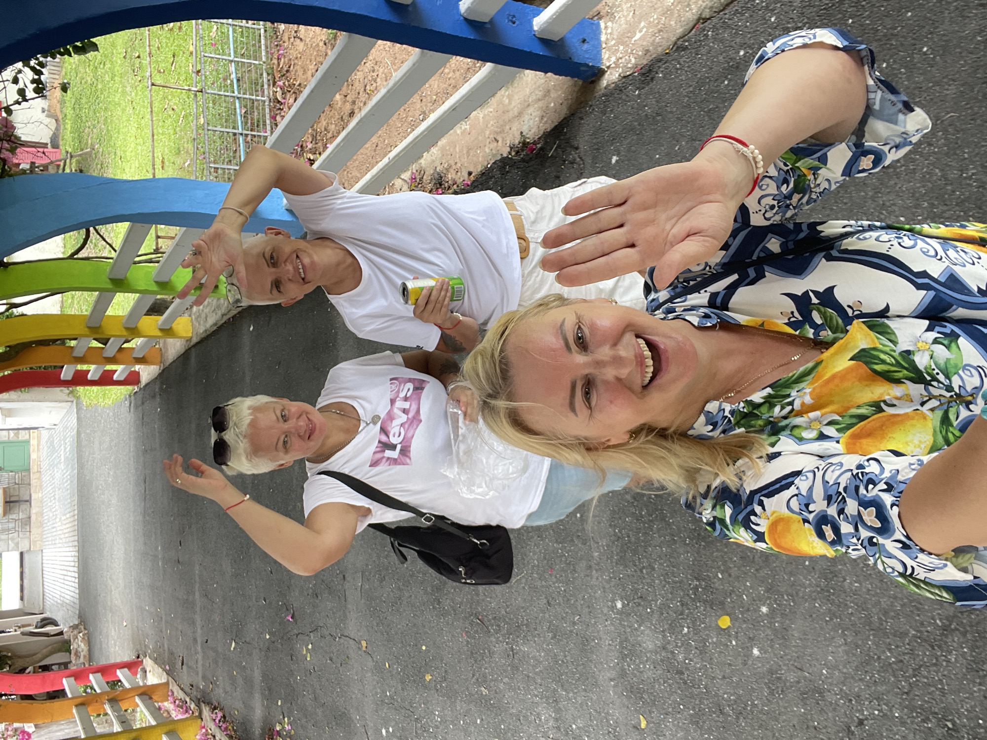Three women are posing for a selfie in an outdoor setting in Na Chom Thian, Thailand. The woman in the foreground is wearing a colorful patterned shirt with lemons and blue designs and is waving at the camera. The other two women are standing behind her. The woman on the left has blonde hair and is wearing a white t-shirt with a pink Levi's logo. She is also wearing sunglasses on her head and has her hand raised. The woman on the right has short blonde hair and is wearing a white t-shirt and white shorts. She is holding a can of drink and has her hand raised as well. They are standing on a paved pathway with colorful arched structures on either side. The background shows greenery and a white fence.