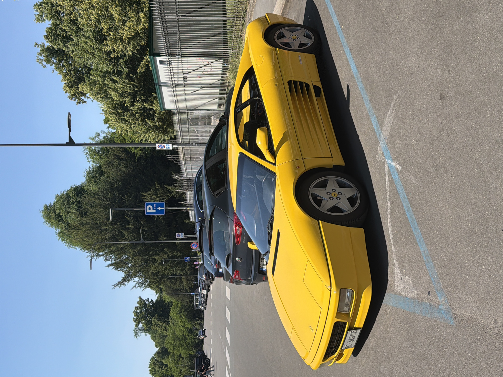 The image shows a yellow Ferrari parked in a designated parking space on a street in Milan, Italy. Other cars are parked behind the Ferrari. Trees and a clear blue sky are visible in the background. There are lampposts and road signs alongside the street. Some people are visible in the distance.