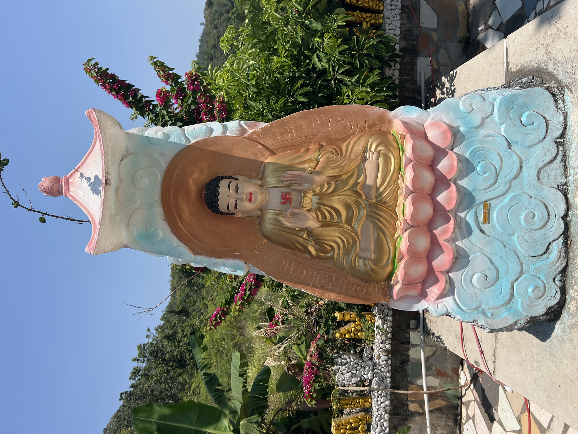 A statue of a seated Buddha with a swastika on the chest, set amidst vibrant foliage, stands in Nha Trang, Vietnam. The statue is painted in gold, peach, and blue hues, with the swastika symbol in red. It is placed against a backdrop of lush greenery and a clear blue sky. The statue is surrounded by flowering plants and a mosaic-style wall.