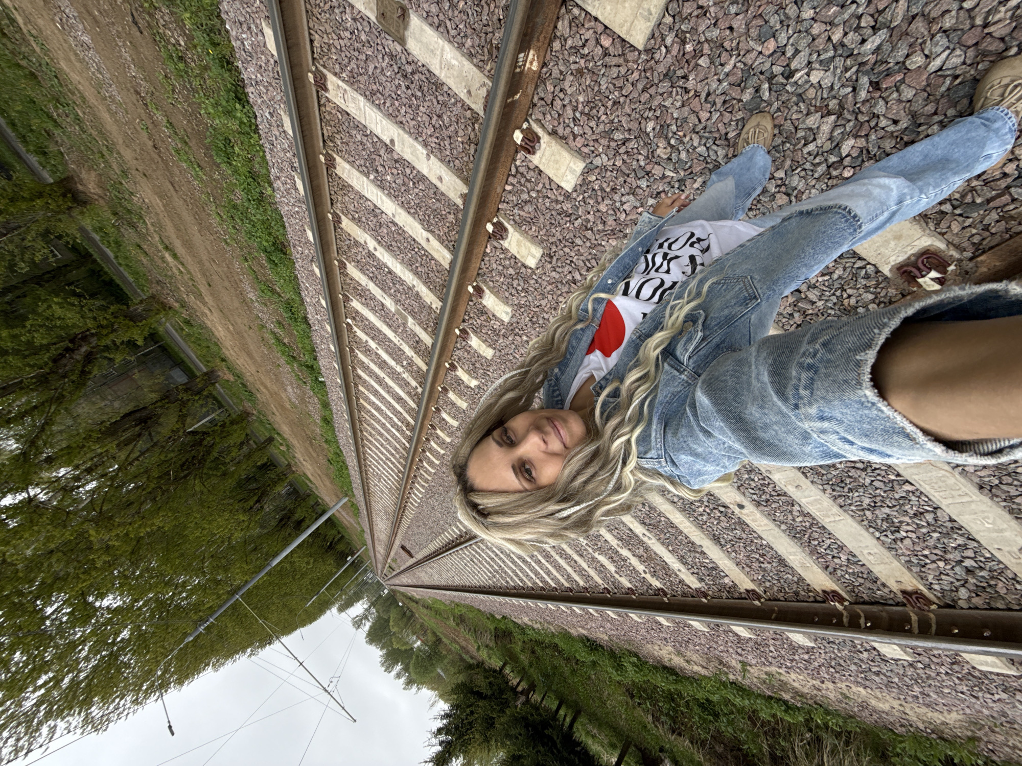 A blonde woman lies on a railway track, smiling at the camera. She is wearing a denim jacket, a t-shirt with writing and a red heart, and jeans. The tracks run into the distance, flanked by gravel and wooden sleepers. On either side of the track is green vegetation and trees. The photograph appears to have been taken in a rural area. There is no confirmation that the location is Rusanovo, Russia.