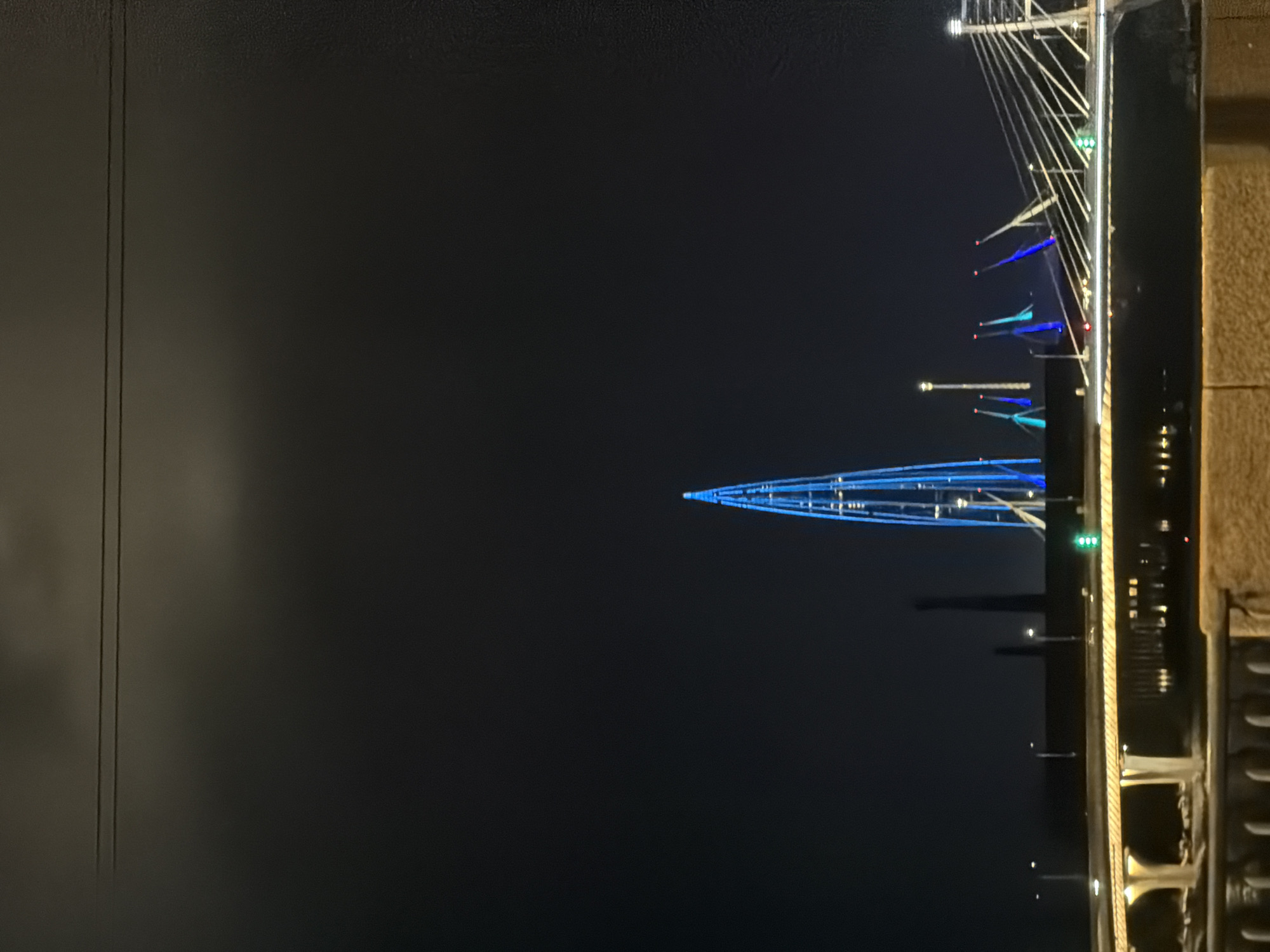 The image captures a nighttime view of the Neva River in Saint Petersburg, Russia, featuring the Neva Palace and the Neva Tower. The Neva Tower, a prominent skyscraper, is illuminated with striking blue lights, creating a vibrant, modern silhouette against the dark sky. In the foreground, a bridge, likely the Big Obukhovsky Bridge, is visible with its deck illuminated by warm, yellow lights, and suspension cables stretching across the frame. The river reflects the city lights, adding depth to the scene. The sky is dark with faint clouds, suggesting a clear night. The overall atmosphere is one of urban beauty and modern architecture harmonizing with historical surroundings.
