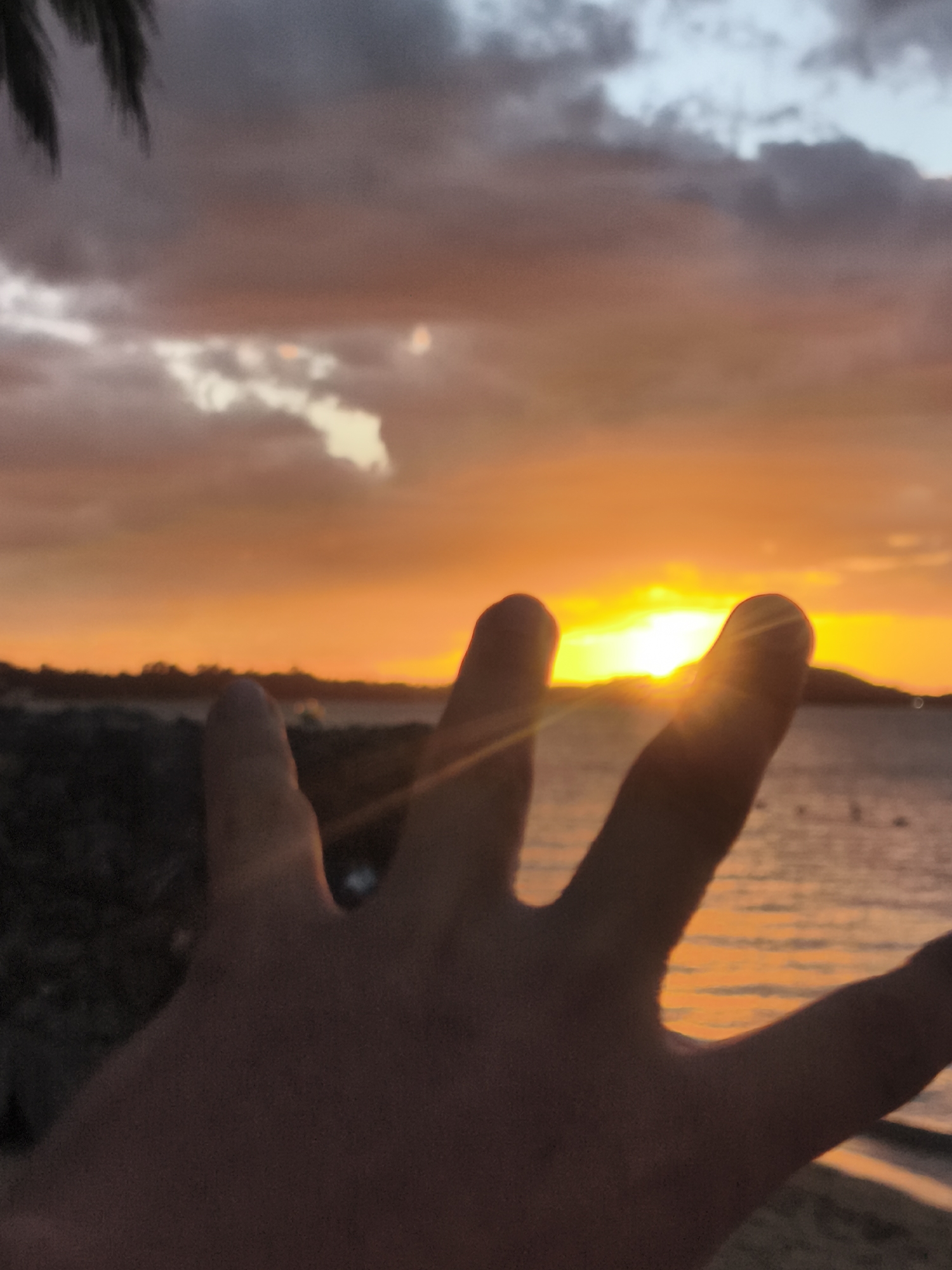 A close-up, over-the-shoulder shot shows a hand with fingers spread in the foreground, obscuring a sunset over the ocean. The sun is visible as a bright orb behind the fingers, casting orange and yellow light across the sky. The ocean is calm with gentle waves reflecting the sunset colors. In the distance, a silhouetted coastline with trees and hills is visible. The sky is filled with dramatic clouds in shades of orange, pink, and gray. A palm frond is visible in the upper left corner of the frame. The scene appears to be on a beach in Baan Bo Phut, Thailand, likely during a vacation.