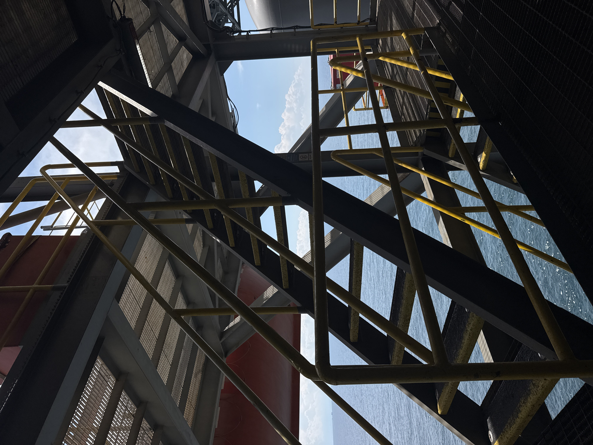 A close-up, low-angle, outdoor shot captures a metal staircase with yellow handrails on what appears to be an industrial platform, possibly an offshore oil rig or ship, with the ocean and sky visible in the background. The staircase is constructed from dark metal with yellow-painted railings and steps. The grating of the walkway and the underside of another deck are visible in the foreground and upper portions of the image, respectively. The sea is a deep blue, and a few white clouds are scattered across a bright blue sky. There are no people or animals visible in the frame. The overall impression is one of functional, industrial infrastructure against a natural backdrop. The location is not identifiable as being in any specific city in Italy.
