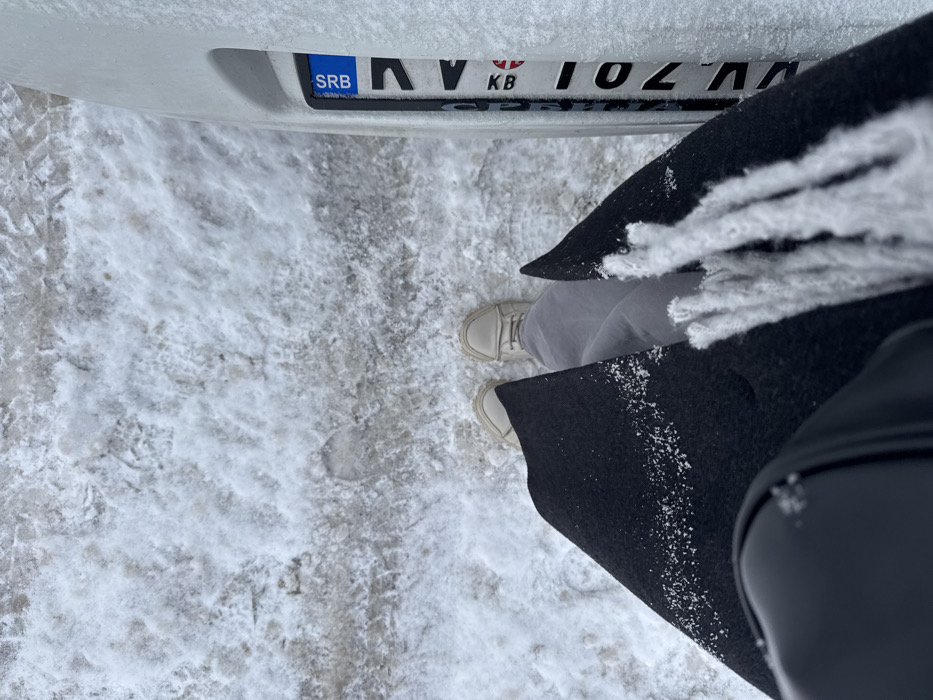 An overhead view displays a person standing on snow and slush-covered ground in Cibukovac, Serbia. The individual wears light-colored athletic shoes, grey pants, and a dark coat with visible snow specks. A dark bag is positioned on the left side of the coat, and a white fringed textile, possibly a scarf, is draped over the right side of the coat. To the right of the person, the rear section of a white vehicle is partially visible, featuring a Serbian license plate. The license plate displays the "SRB" country code and characters including "AV", "KB", and "701", with the plate itself partially covered by snow and ice. Tire tracks and a distinct footprint are visible in the snow and ice mixture on the ground.