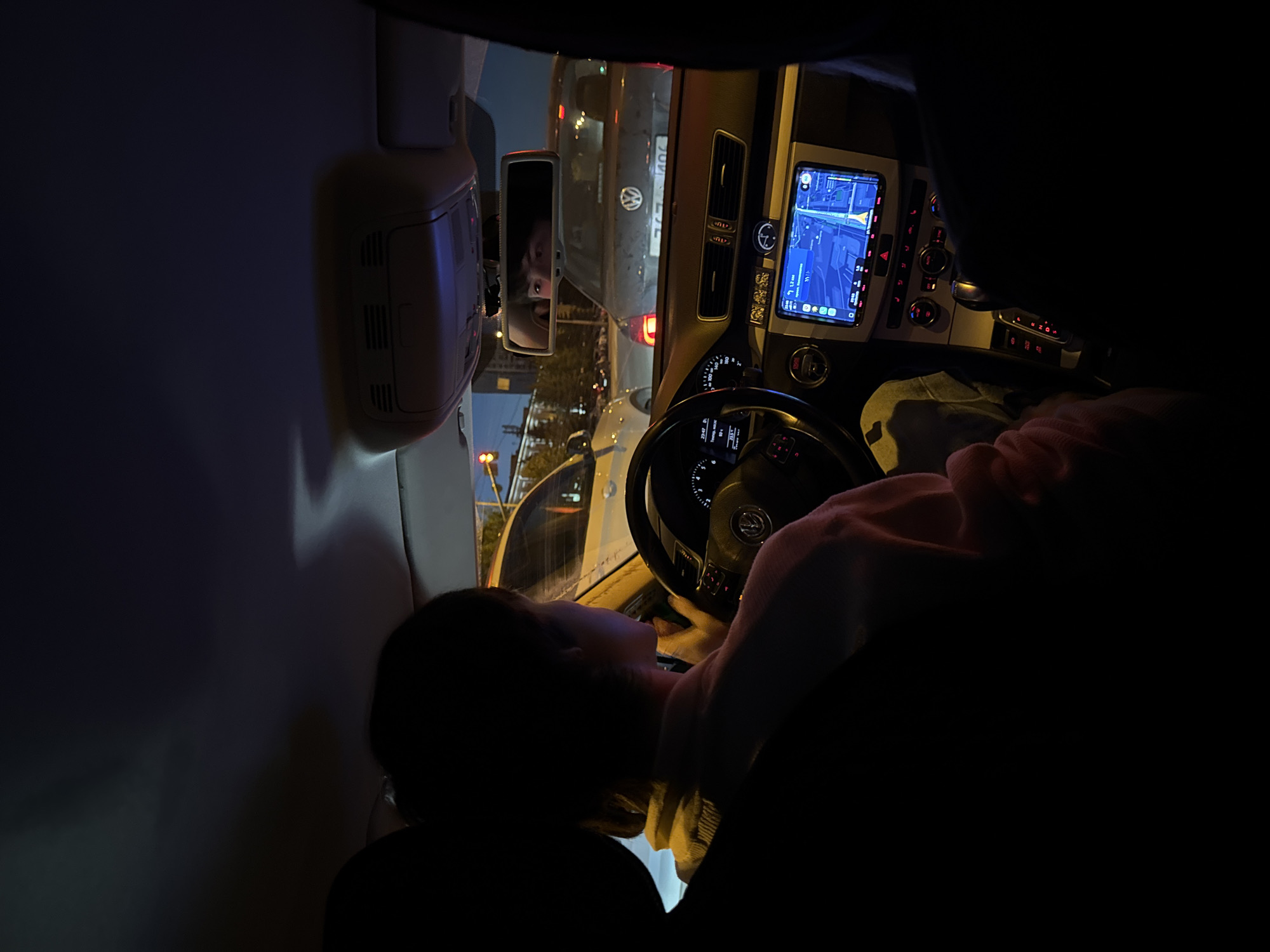 The image is an interior shot of a car at night, possibly in Yekaterinburg, Russia, based on the surrounding context. A person, identified as female by their profile, is in the driver's seat, facing forward. They are wearing a light-colored sweater and have short, dark hair. The car's interior is visible, including the dashboard with a navigation screen displaying a map, the steering wheel with a Volkswagen logo, and the gear shift. The car appears to be stopped, possibly in traffic, as evidenced by the vehicle in front of it with its brake lights illuminated. The lighting suggests it is nighttime, with artificial lights from streetlights and other vehicles illuminating the scene. The reflection in the rearview mirror shows another person, likely the photographer, looking at the camera. The overall atmosphere is dark and intimate, capturing a moment of travel within the city.