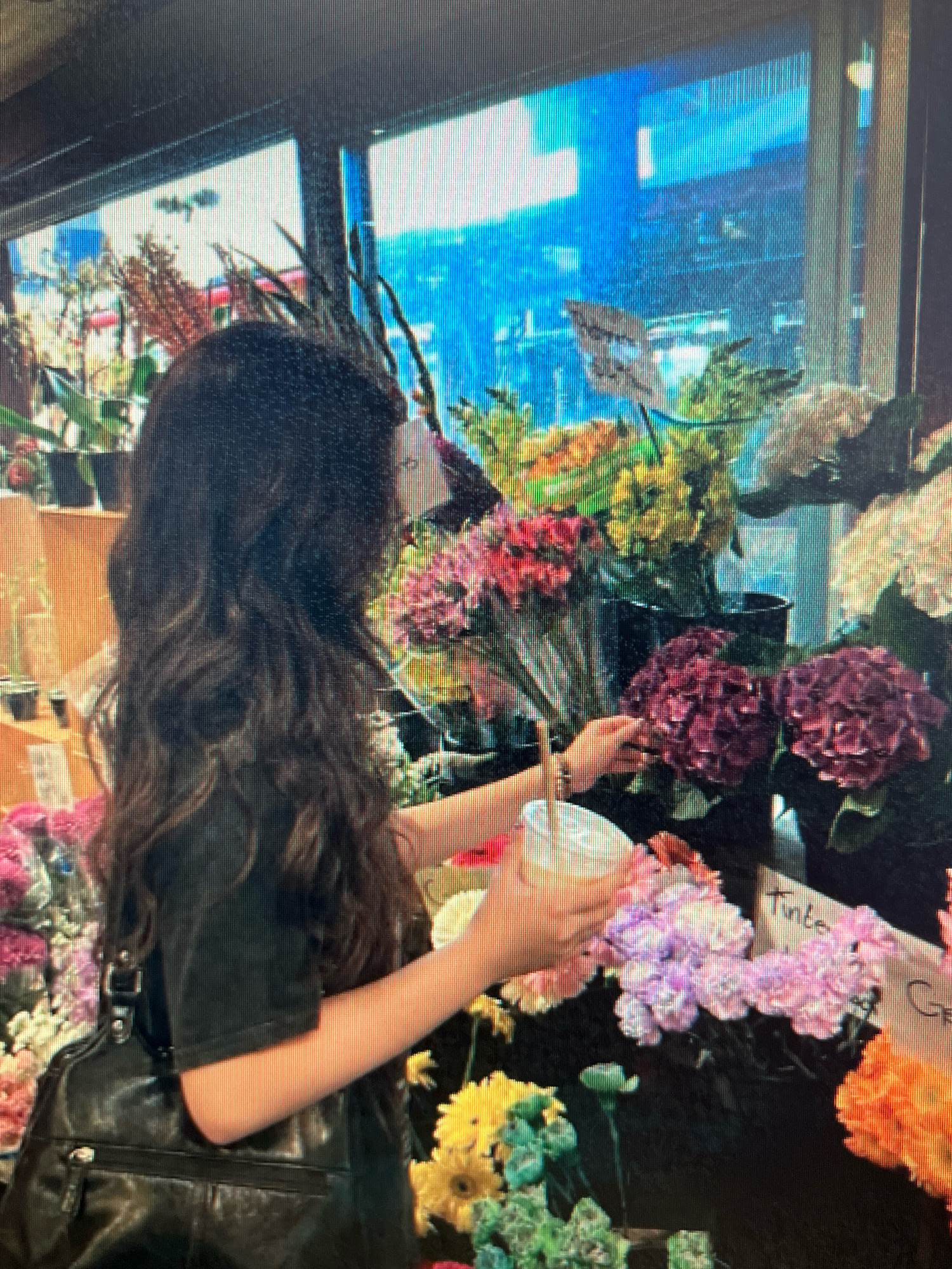 The image shows a person with long, dark, wavy hair, seen from the back, standing in front of a flower stall. The person is wearing a dark top and carrying a black purse. They are holding a light-colored cup in their right hand and appear to be selecting flowers. The stall is filled with a variety of colorful flowers, including pink, purple, yellow, and orange varieties. In the background, through a window or glass, a blue and bright urban scene is visible, possibly indicating a street or a building. The lighting suggests it is daytime. The location is the city of Sejahtera, Indonesia.
