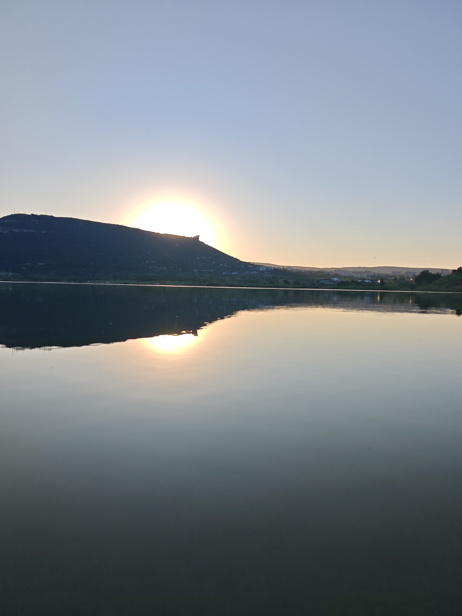 The image captures a serene sunset over the Vreteno Dam, located in the city of Shumen, Bulgaria. The sun, a brilliant orb of light, is partially obscured by the silhouette of a large, dark hill on the left side of the frame. This hill, characteristic of the Shumen Plateau, is dotted with a few structures, possibly communication towers, visible against the bright sky. The sun's reflection creates a shimmering path of light across the calm surface of the dam, which occupies the lower half of the image. In the distance, along the horizon, a town or city is visible, its lights twinkling faintly. The sky is a soft gradient of blue, transitioning to a lighter hue near the horizon. The overall atmosphere is tranquil and picturesque, showcasing the natural beauty of the area at dusk. There are no people visible in the image.