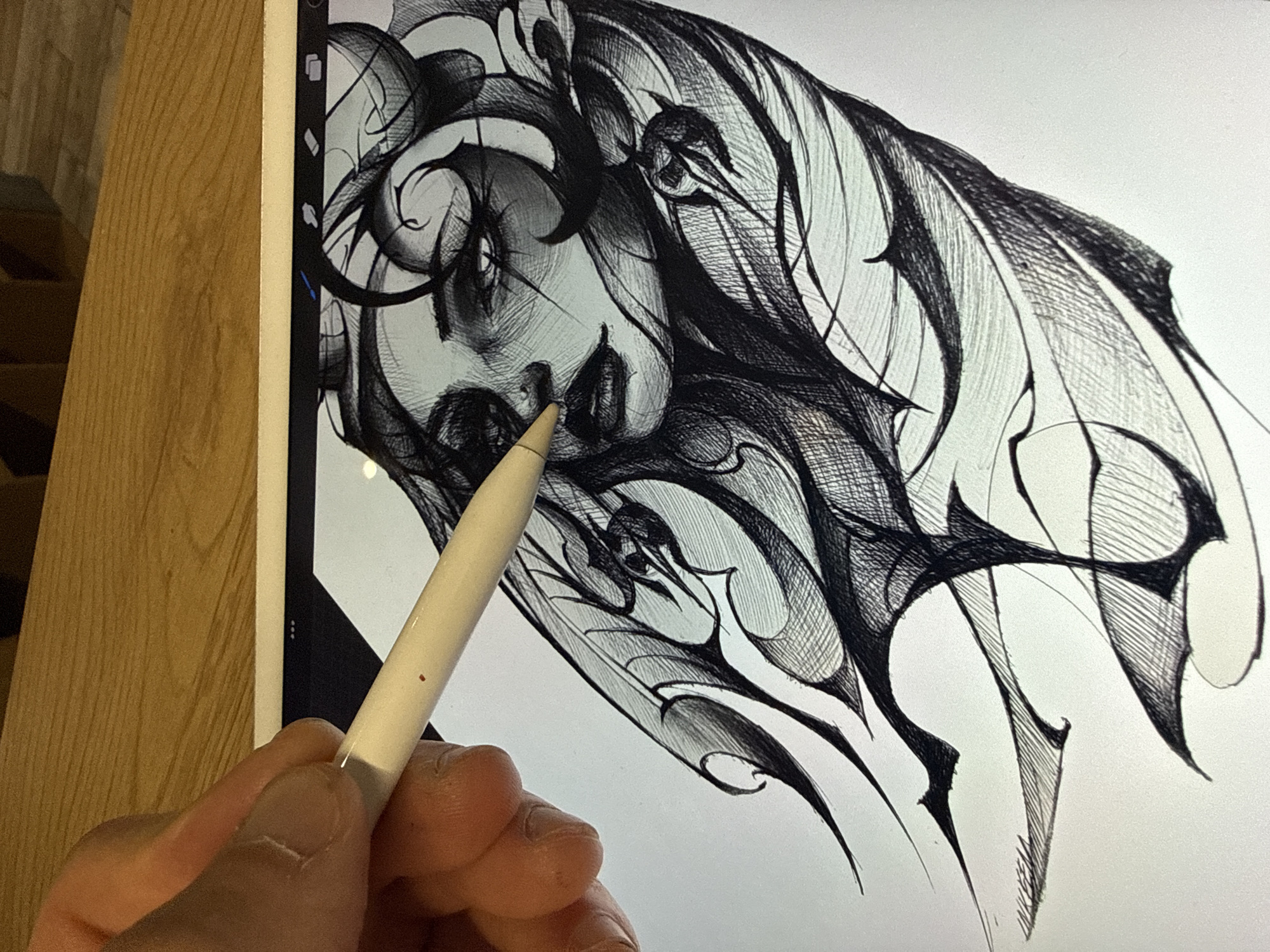 The image shows a close-up of a person's hand holding a white digital stylus, actively working on a drawing on a tablet screen. The stylus, likely an Apple Pencil, is positioned near the center of the digital artwork.
The artwork displayed on the tablet is an intricate, black and white illustration. It features a stylized human face, possibly female, with expressive, dark eyes, partially obscured and integrated into a complex design of swirling, organic, and abstract elements. The drawing employs fine lines and cross-hatching techniques to create texture, depth, and a detailed, somewhat gothic or tribal aesthetic. The style merges portraiture with decorative, flowing forms.
The tablet, appearing to be an iPad, shows a thin bezel and a sliver of its user interface on the left side, though specific icons are not clearly discernible. The artist's hand is visible on the lower right, holding the stylus in a natural grip, with visible knuckles and fingernails. A small red dot is noticeable on the side of the stylus.
To the left of the tablet, a light-colored wooden surface with a distinct grain, possibly a desk or tabletop, occupies a significant portion of the background. A darker, textured element is partially visible in the upper left background, behind the wooden surface. The scene is illuminated by ambient indoor light. The activity depicts a moment of creative focus in a digital art workflow, taking place in Sofia, Bulgaria. No explicit text is visible in the image.