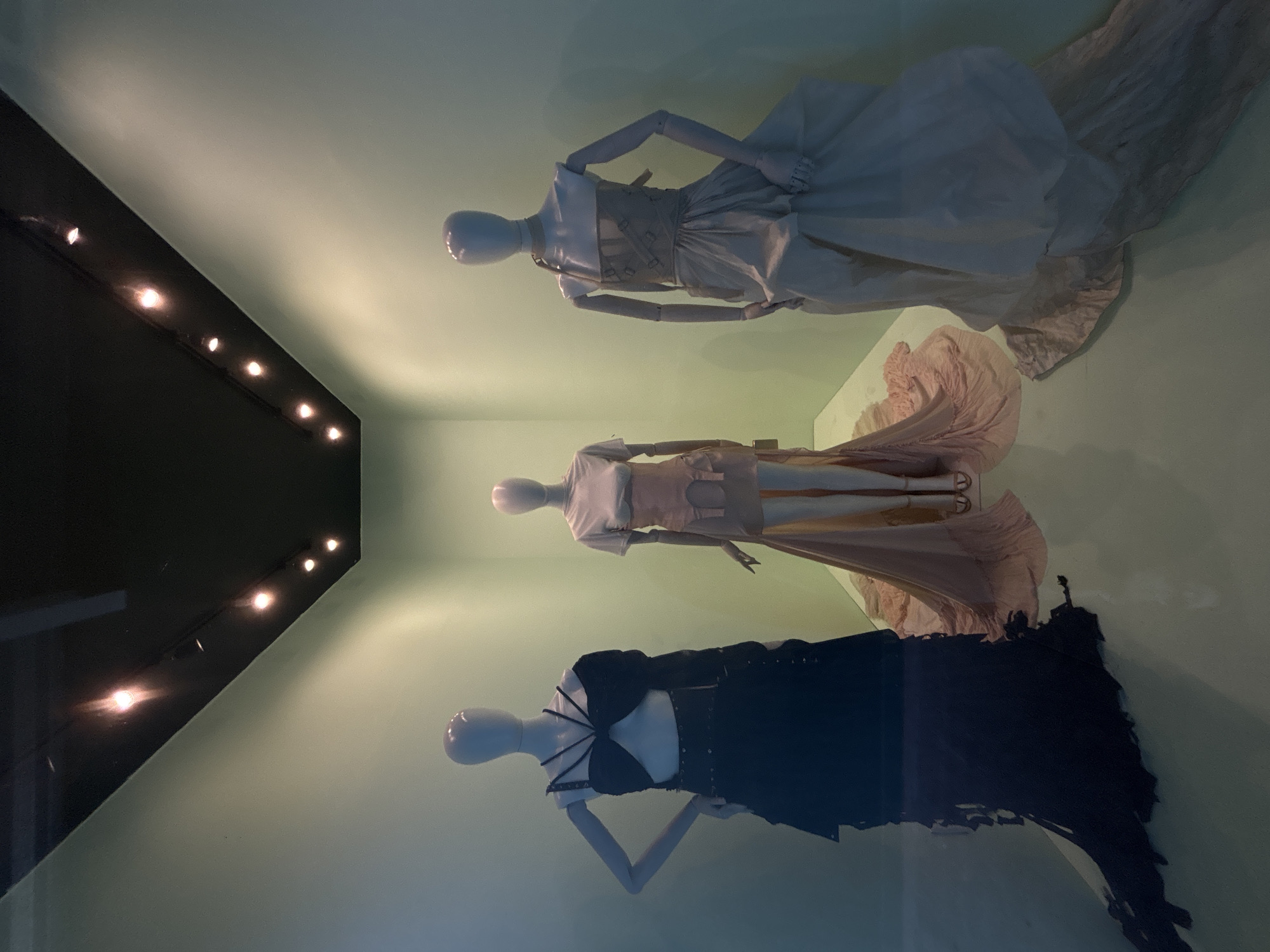 This media file captures an indoor exhibition scene featuring three mannequins displaying elaborate fashion garments, likely within a museum or gallery setting in Mexico City, Mexico. The image appears to have been taken with the camera rotated, as the mannequins are oriented horizontally but are depicted as if standing upright.
The three full-body mannequins are positioned against a muted, light green wall. From left to right:
1. The first mannequin wears a dark, possibly black, two-piece ensemble. The top is a distinctive bralette-style or cropped piece with multiple straps converging on the chest, paired with a long, voluminous skirt that exhibits a textured or layered appearance.
2. The middle mannequin displays a pale pink or beige two-piece outfit. This includes a fitted, short-sleeved top with geometric cut-outs along the sides and a flowing, long skirt with a train.
3. The third mannequin is dressed in a light-colored, possibly off-white or cream, two-piece gown. The top features a structured, corset-like bodice with a high neck and straps, while the skirt is voluminous and floor-length with a long, trailing train.
All mannequins have smooth, featureless heads and articulated, light-colored wooden arms, suggesting a stylistic choice for the display.
A row of warm, bright spotlights illuminates the scene from the left, running along an angled dark surface that could be part of the display architecture or ceiling. A prominent reflection of these lights and parts of the room is visible across the foreground, indicating the display is behind a glass panel or reflective barrier. The lighting is controlled and dramatic, typical of an exhibition space, making the time of day undeterminable. No visible text is present in the image.