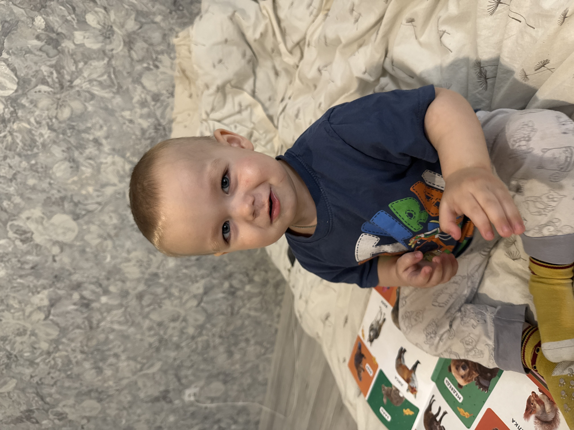 The image, rotated 90 degrees clockwise, features a young toddler sitting or lying on a blanket indoors. The child, with light blonde hair and blue eyes, looks directly at the camera with a broad, happy smile. They are dressed in a navy blue long-sleeved t-shirt adorned with colorful, cartoon-like animal or character appliques, light-colored patterned pants, and yellow and grey striped socks with anti-slip dots.
The child's hands are resting near an open children's book, which features illustrations of animals on its pages. Clearly visible animals include a fox, a bear, and a squirrel. Accompanying text in Cyrillic script identifies the animals: "ЛИСА" (Fox), "МЕДВЕДЬ" (Bear), and "БЕЛКА" (Squirrel).
The setting appears to be a cozy indoor environment, likely a bedroom or play area. The child is on a cream or white blanket patterned with delicate, dandelion-like seed heads. Behind the child and blanket, a wall covered in grey or silver wallpaper with an ornate floral or abstract design is visible. The lighting suggests it is daytime. The child's expression conveys happiness and engagement, possibly with the book.