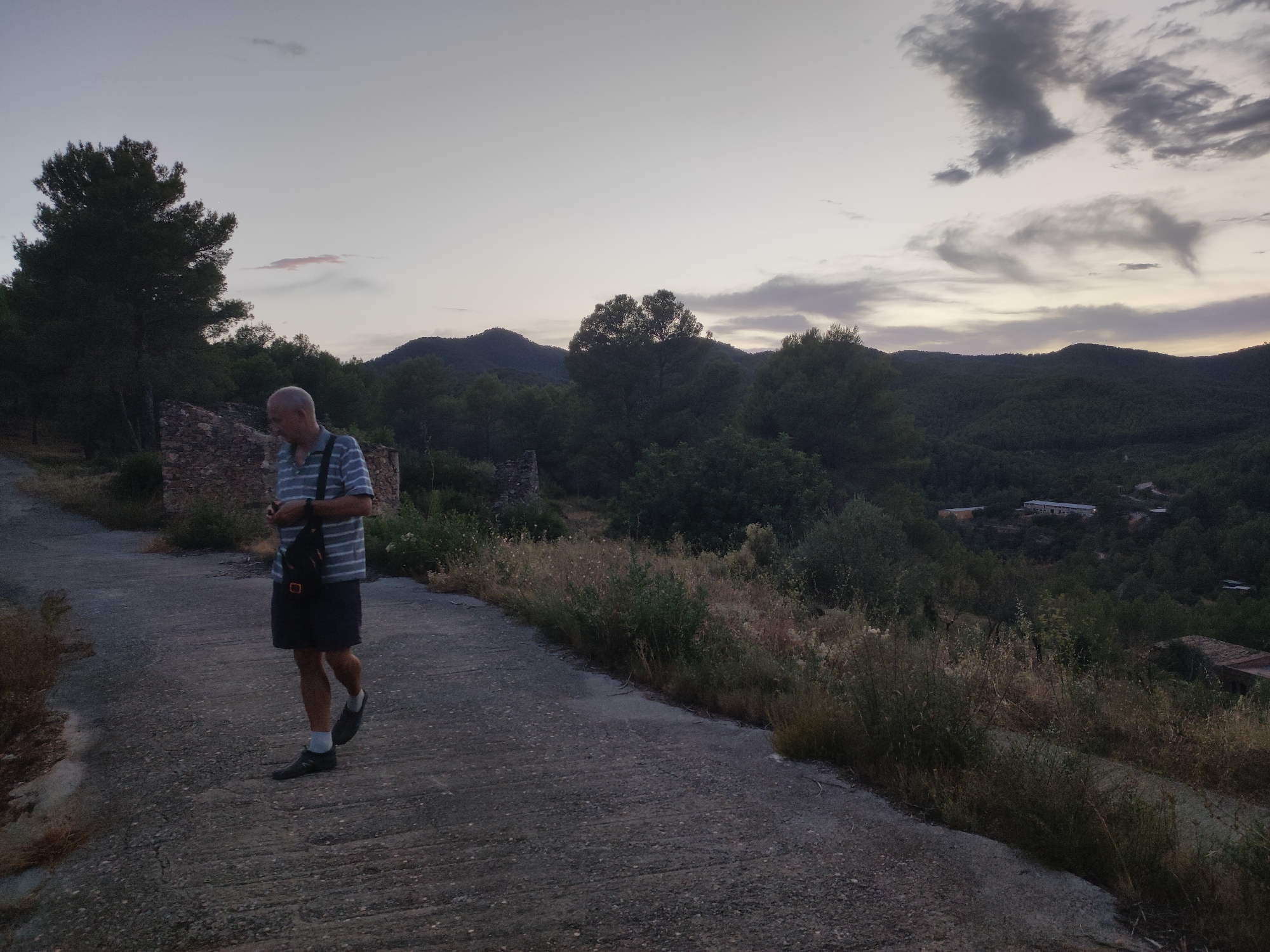 An elderly man with graying hair, wearing a striped t-shirt, dark shorts, and black shoes with white socks, walks along a paved road. He is carrying a dark bag slung across his chest. The road is surrounded by dry vegetation and a stone wall. In the background, rolling hills covered in trees and vegetation extend to the horizon, under a pale sky with wispy clouds. Several buildings are visible nestled in the hills. The scene appears to be at dusk. The location is the municipality of Suera/Sueras, Spain, within a rural, hilly landscape.