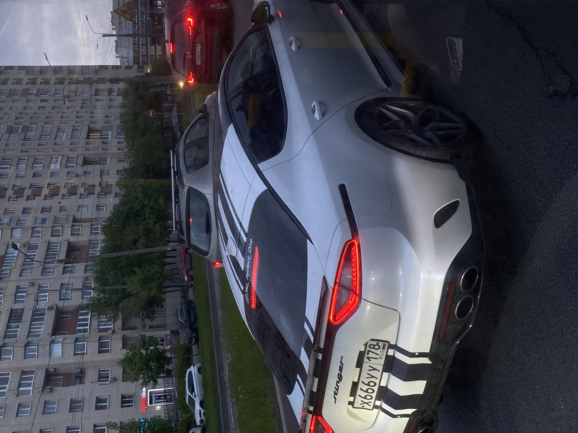 The image shows a white Kia Stinger parked on a street in Saint Petersburg, Russia. The car has black and white stripes on the rear and the license plate reads "x666yy 178 RUS". The car is parked facing away from the camera. In the background, there are apartment buildings, trees, and other vehicles, including a red Mazda. The sky is overcast.