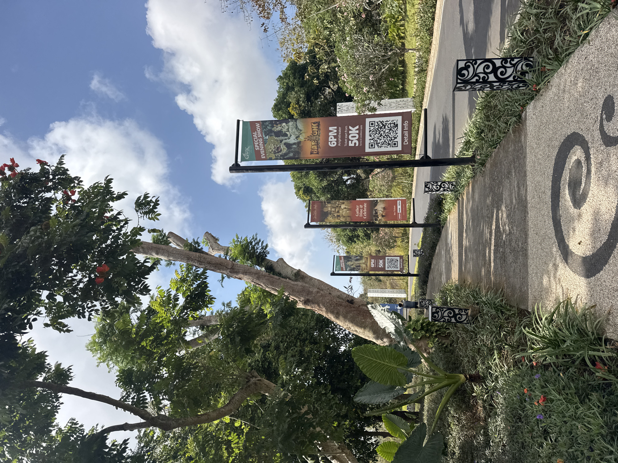 A paved walkway curves along a road in Kuta Selatan, Indonesia, bordered by lush greenery and large trees. Several tall, vertical banners are posted along the roadside. One banner advertises a "SPECIAL EVENING SHOW" featuring "BARONG" at 6 PM in the Amphitheater, with tickets priced at 50K, and includes a QR code for more information. Another banner, to the left, advertises "Gather, Dine & Celebrate" with operating hours from 9 AM to 10 PM. Ornate, black metal lanterns line the walkway. The sky is blue with scattered white clouds.
