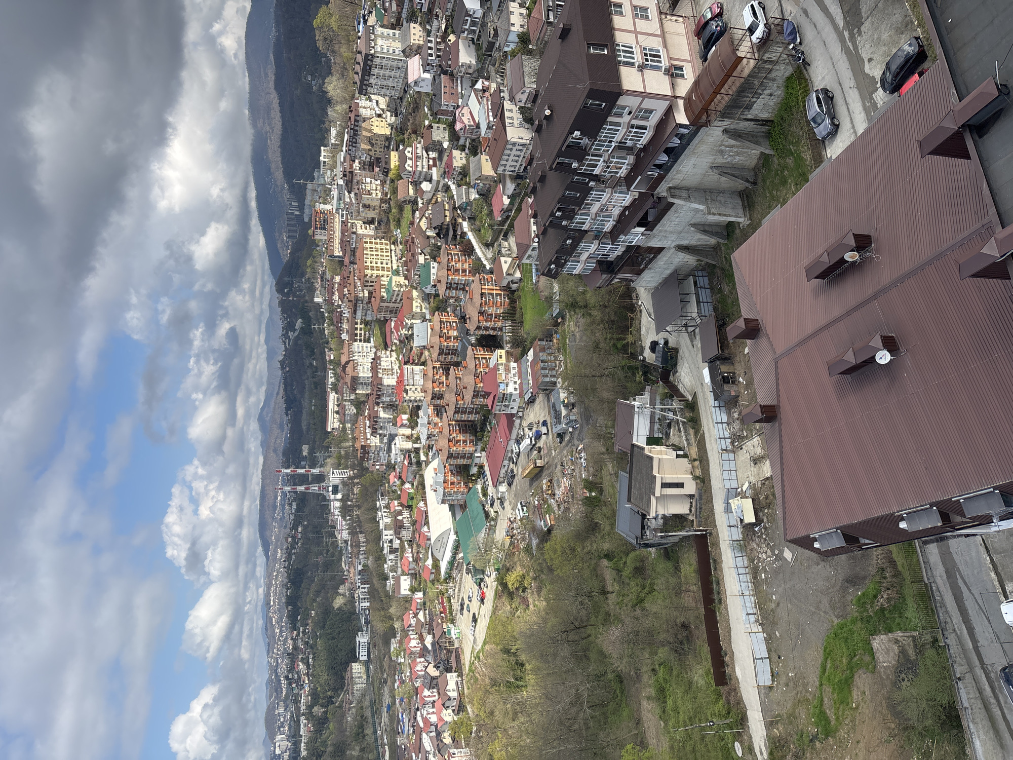 The image presents an elevated view of a city nestled in a valley, likely Sochi, Russia, given its mountainous backdrop and architecture. The foreground includes the brown roof of a multi-story building, viewed from above. Adjacent to the building is a paved area with some parked cars.
Beyond the immediate foreground, the cityscape unfolds with closely packed buildings, a mix of residential and commercial structures. The buildings have varied roof colors, including red, brown, and green. The city extends towards the base of the mountains in the distance.
The background is dominated by mountains partially obscured by clouds, with patches of blue sky peeking through. The sky is filled with cumulus clouds.