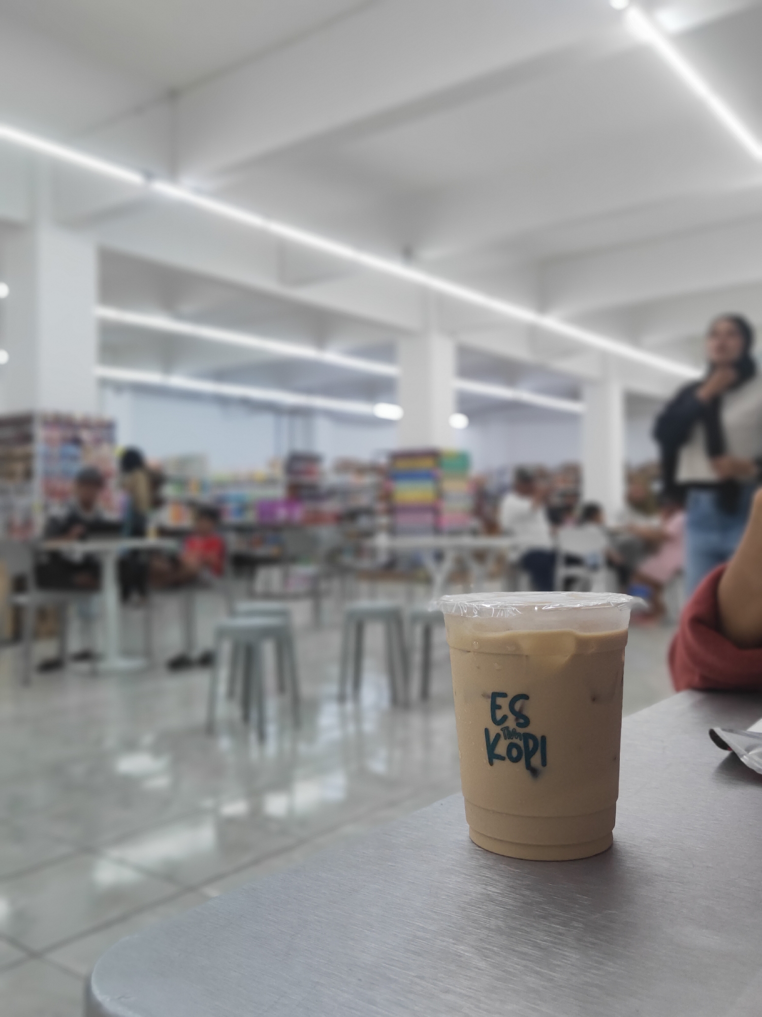 The image captures a close-up of an iced coffee cup with the text "ES KOPI" visible on its side. The cup is placed on a metallic table. The background is blurred, showing an indoor environment with people and shelves filled with products, suggesting a retail setting such as a minimarket or a cafe within a larger establishment. The lighting appears to be artificial, with strip lights visible on the ceiling. The overall scene is casual and everyday.