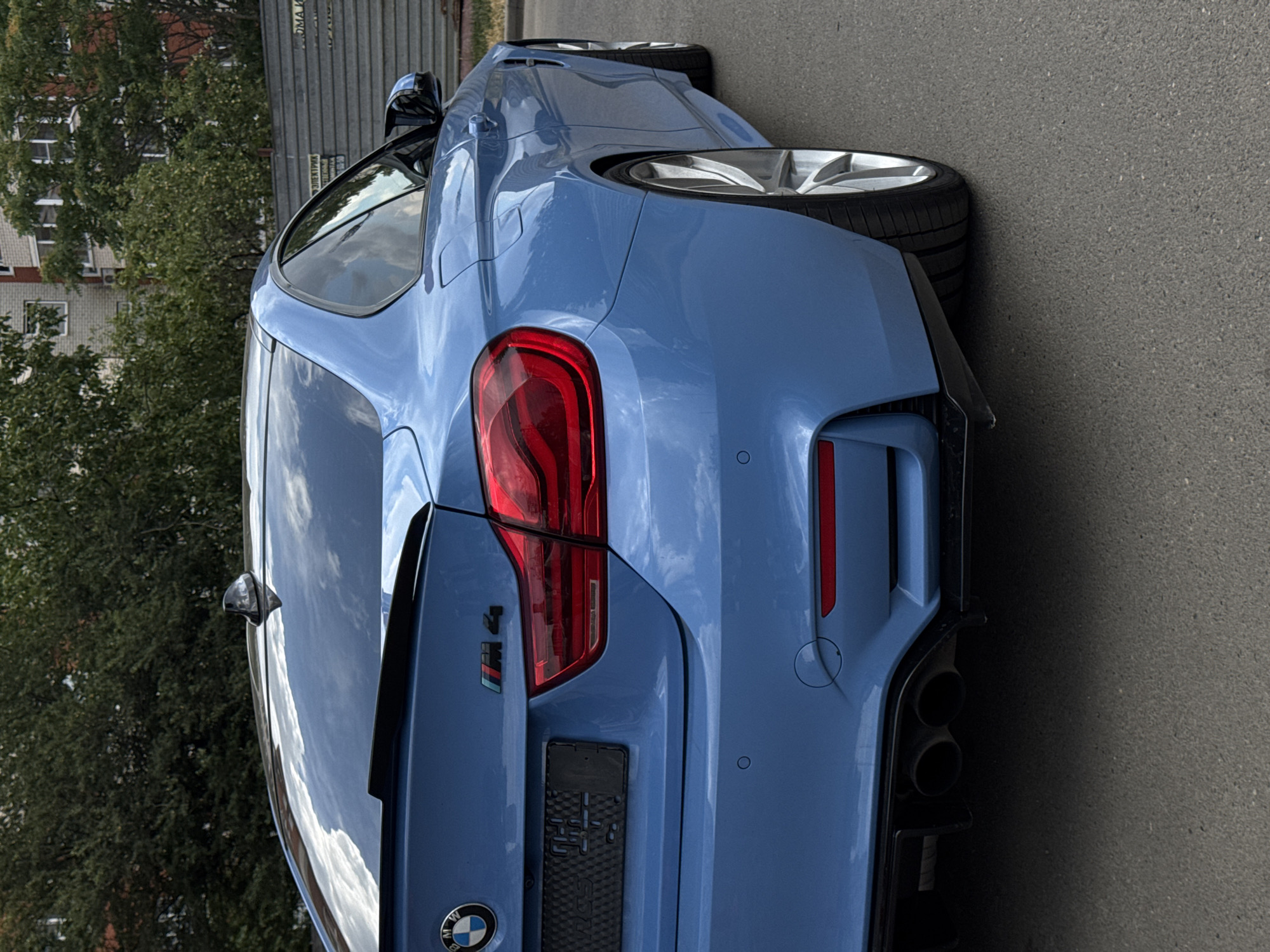 A blue BMW M4 coupe is parked on an asphalt surface in front of a gray metal fence and a brick building, surrounded by green trees. The car is facing right, with its rear end prominently visible. The vehicle has a black spoiler on the trunk, a shark fin antenna on the roof, and distinctive M4 badging. The license plate area is empty. The car's taillights are illuminated red. The background shows residential buildings and lush foliage. The scene appears to be in an urban or suburban setting in Krasnodar, Russia, based on the presence of a sign with Russian text that reads "Электрик" (Electrician) and what seems to be Cyrillic lettering on the license plate frame. No people are present in the image.