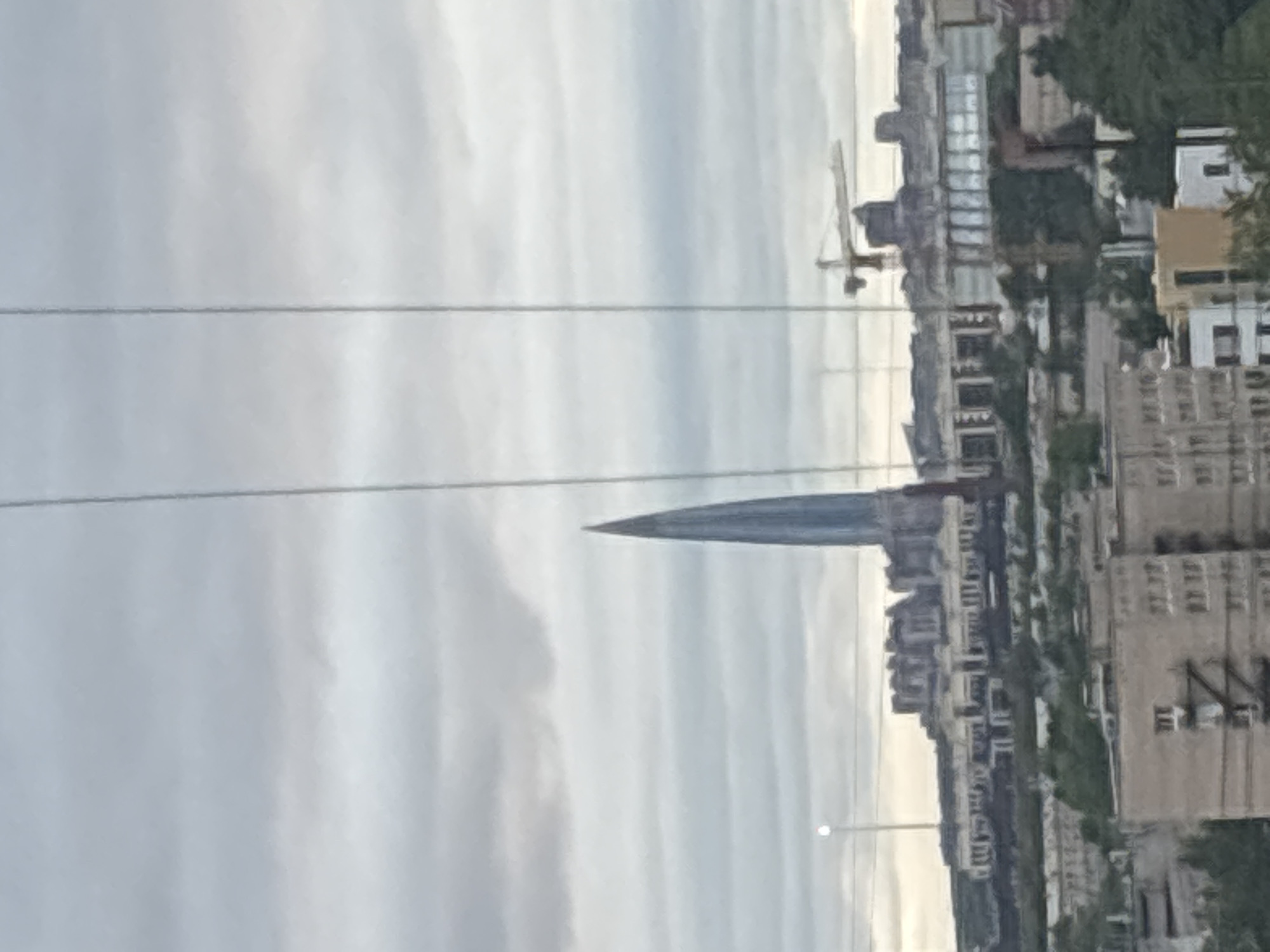 The image depicts a cityscape with a tall, pointed structure dominating the skyline. This structure, possibly a skyscraper or tower, is characterized by its sleek, modern design. In the background, other buildings, trees, and a construction crane can be seen. The sky is overcast, with gray clouds. Power lines are visible in the foreground. The location is within the city of Unknown, Unknown.