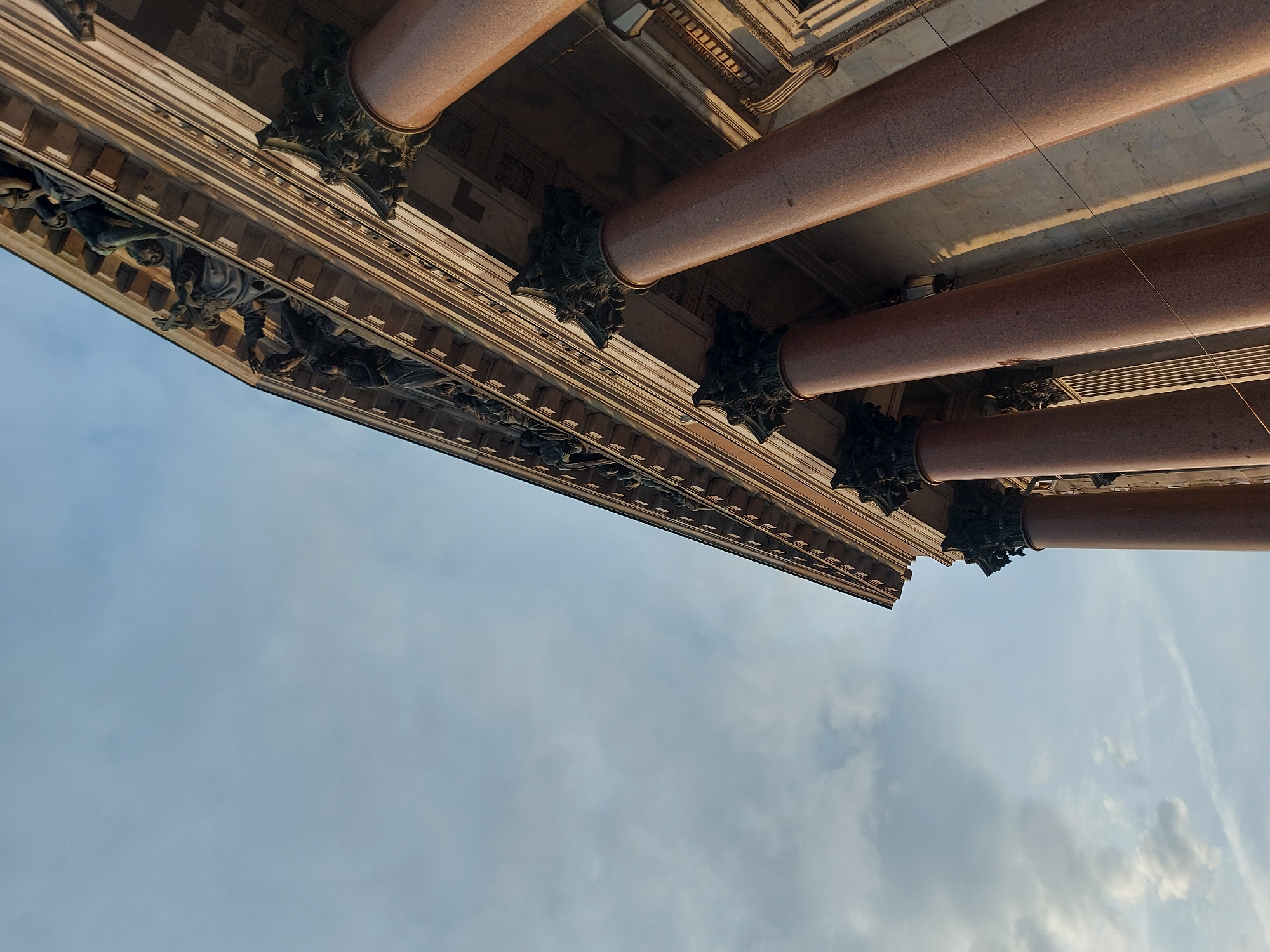 The image shows an upward view of Saint Isaac's Cathedral in Saint Petersburg, Russia. Visible are several large pink granite columns with ornate dark-colored capitals. Below the columns is a detailed entablature adorned with sculptural figures. The background is a sky with light clouds. The angle is low, emphasizing the scale and height of the cathedral.