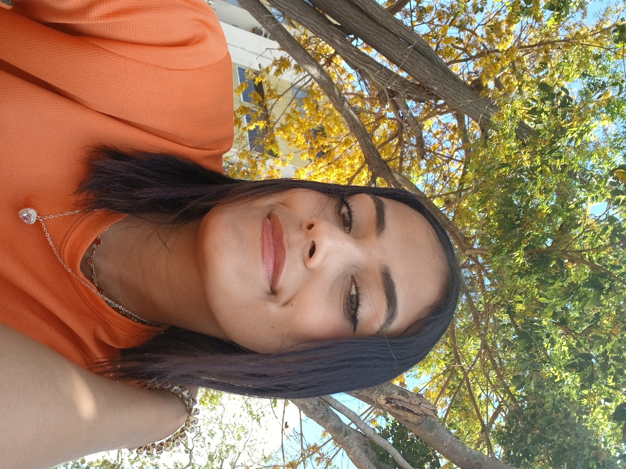 A woman with dark hair and an orange top is pictured in an outdoor setting. The background shows tree branches and foliage with a glimpse of a building. The location is Altagracia, Venezuela.