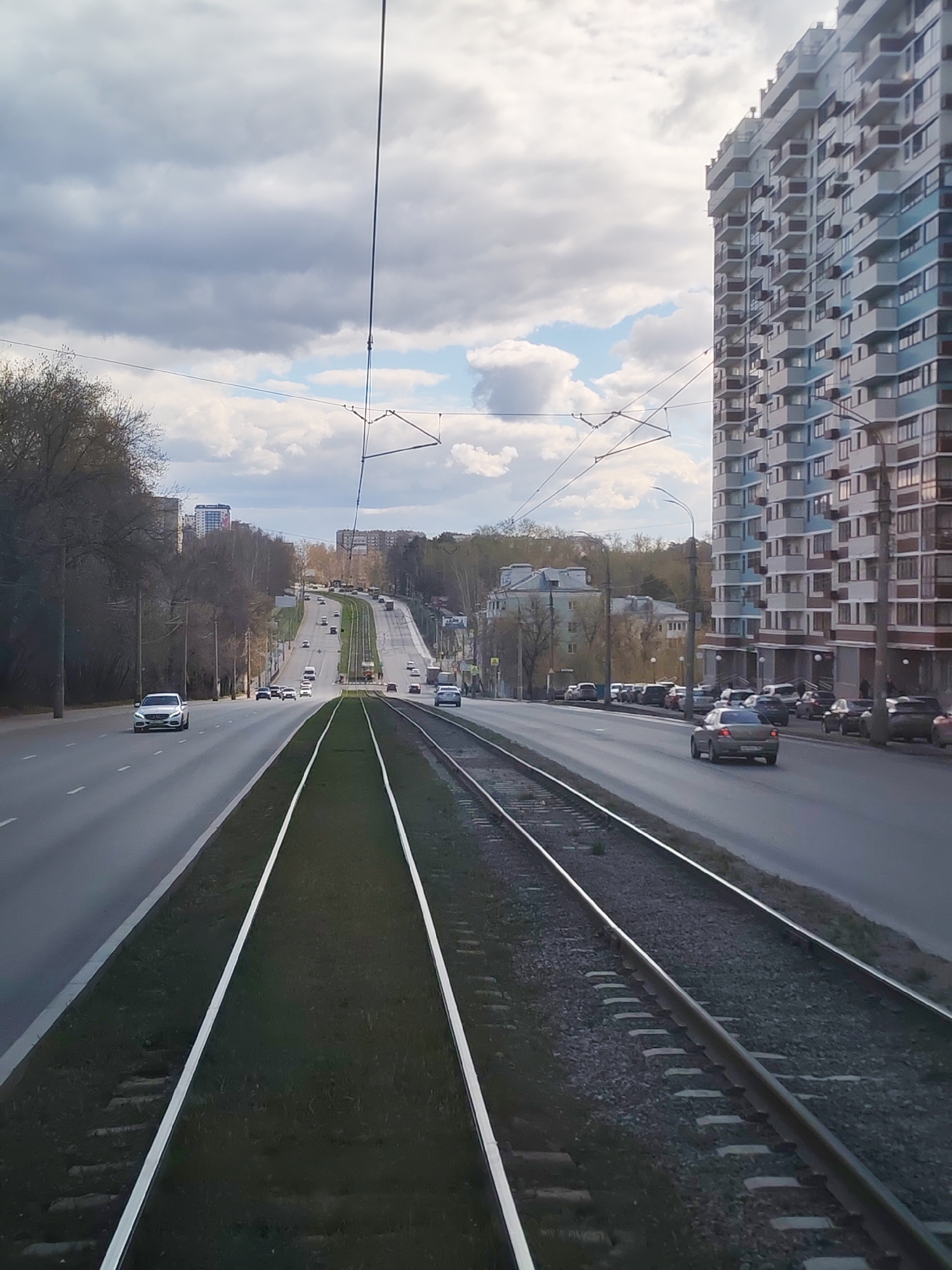 This photo captures a view along a multi-lane street in what appears to be the city of Izhevsk, Russia, notable for its tram tracks running down the center median. The camera angle suggests the photo was taken from within a tram or adjacent to the tracks. A tall apartment building stands prominently to the right of the road, characterized by numerous balconies. Cars are seen driving along the street in both directions. The sky is overcast with patches of blue visible amongst the clouds. There are trees lining the sides of the road. Tram cables are strung overhead.