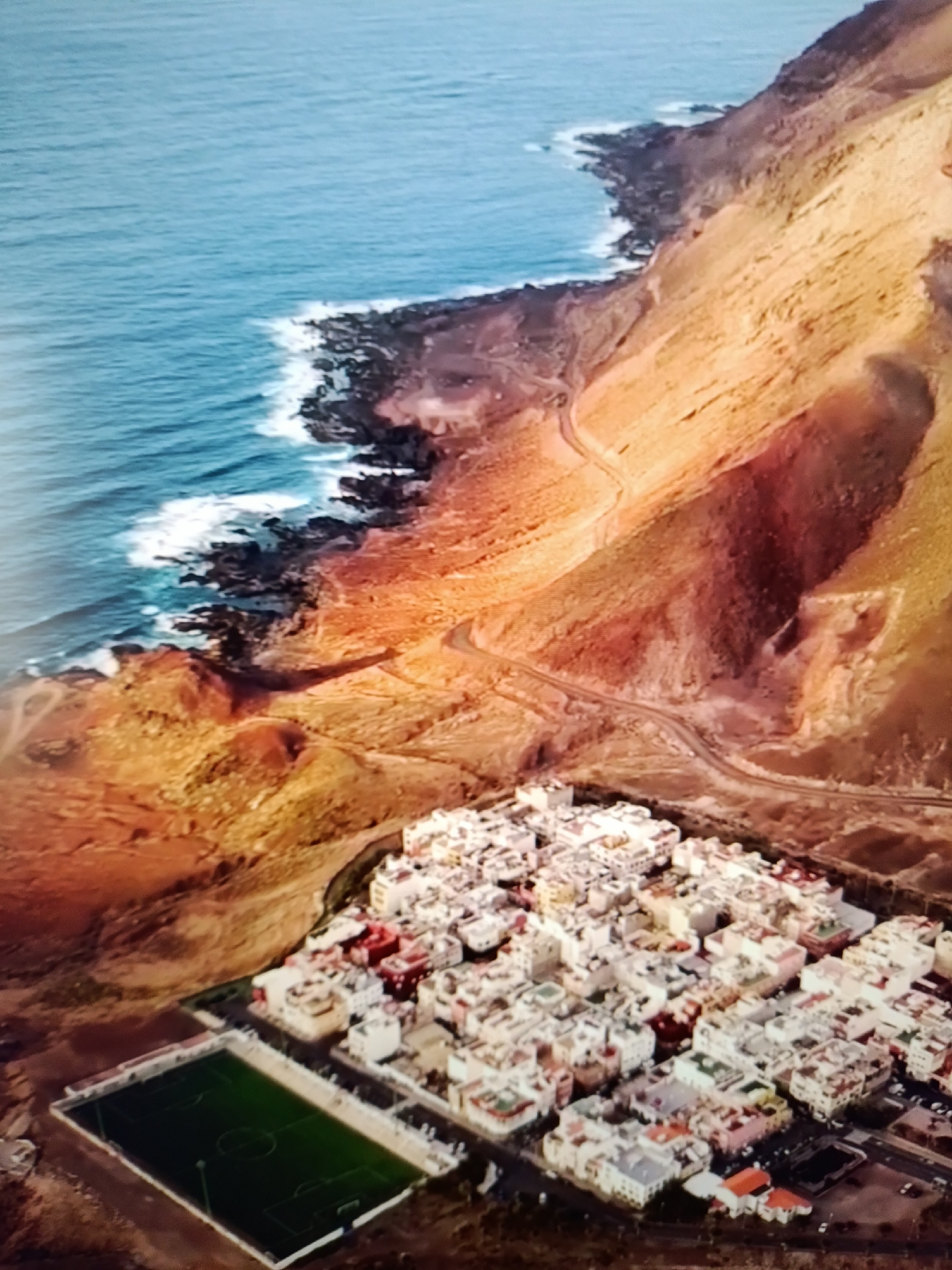 This is an aerial, high-angle, vertical shot of a coastal town. The ocean is on the left with white waves crashing on a rocky, dark-colored shoreline. The land slopes upward from the shore, covered in reddish-brown and orange dirt and vegetation. A winding road snakes its way up the slope. In the lower part of the image, a dense cluster of white and light-colored buildings with red roofs forms a town. Adjacent to the town is a green rectangular sports field, likely a soccer field, with white lines and markings. There are no people clearly visible in the image. The lighting suggests it could be late afternoon or early morning due to the warm tones on the hillside.