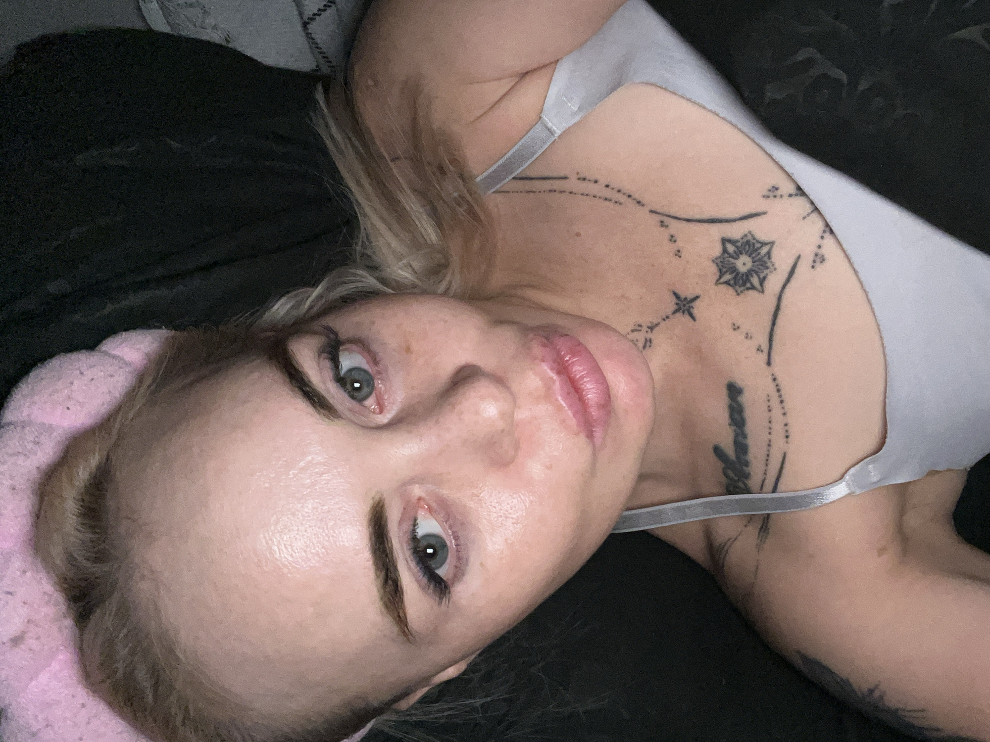 In an overhead shot, a young Caucasian woman with blonde hair, wearing a pink headband, lies on a dark surface. She has blue eyes, visible freckles on her cheeks, and is wearing a light gray top. Tattoos are visible on her chest and shoulder. Her lips are slightly pursed, and she is looking directly at the camera.