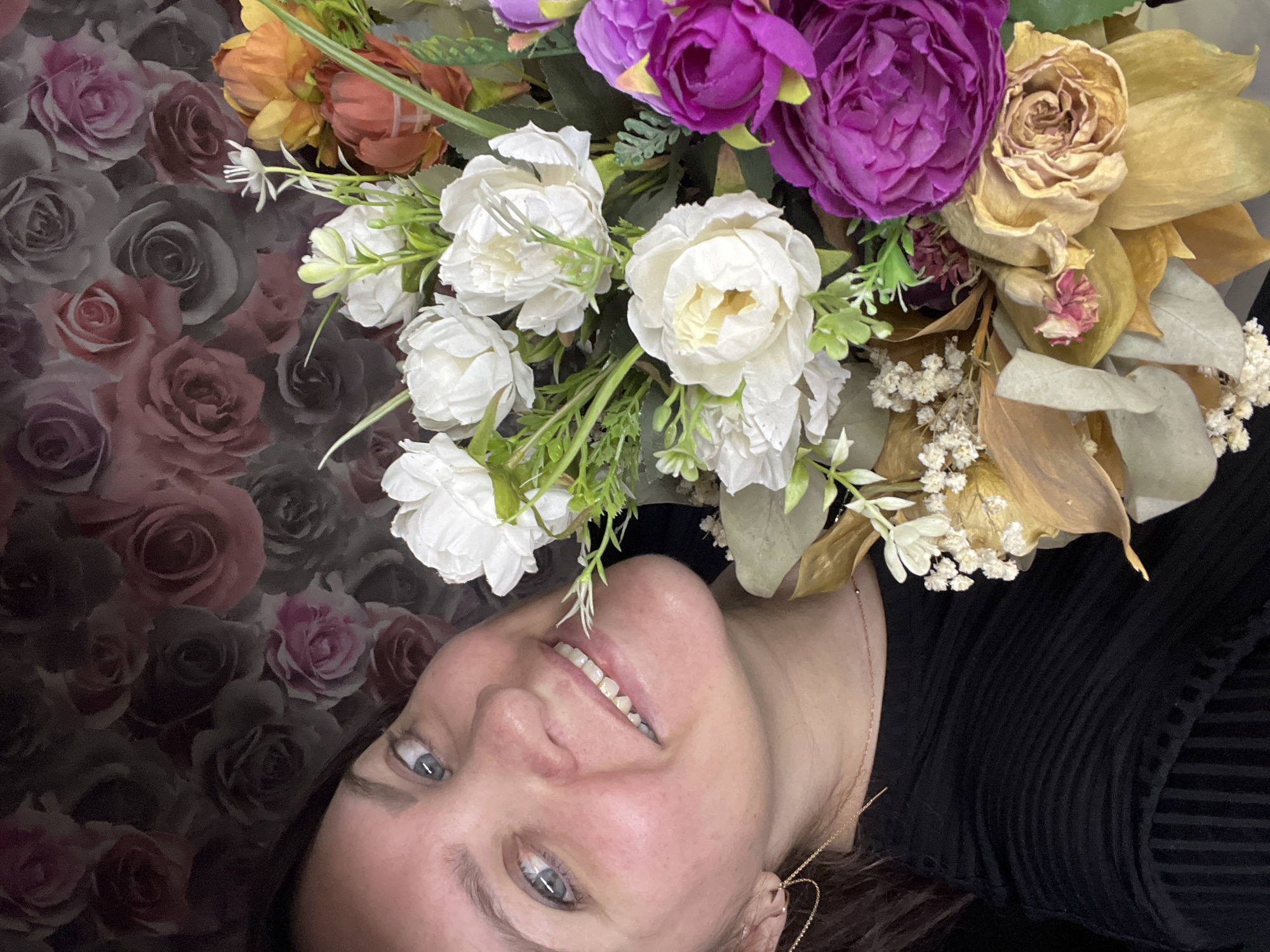 A woman lies on a floral-patterned surface, with a bouquet of flowers positioned above her head. The bouquet contains white, purple, orange, and beige flowers. The woman is wearing a black shirt. A thin gold chain is visible around her neck, and a delicate gold earring is also visible. The woman's eyes are light blue. There is no indication of the location being in Noviy, Russia.