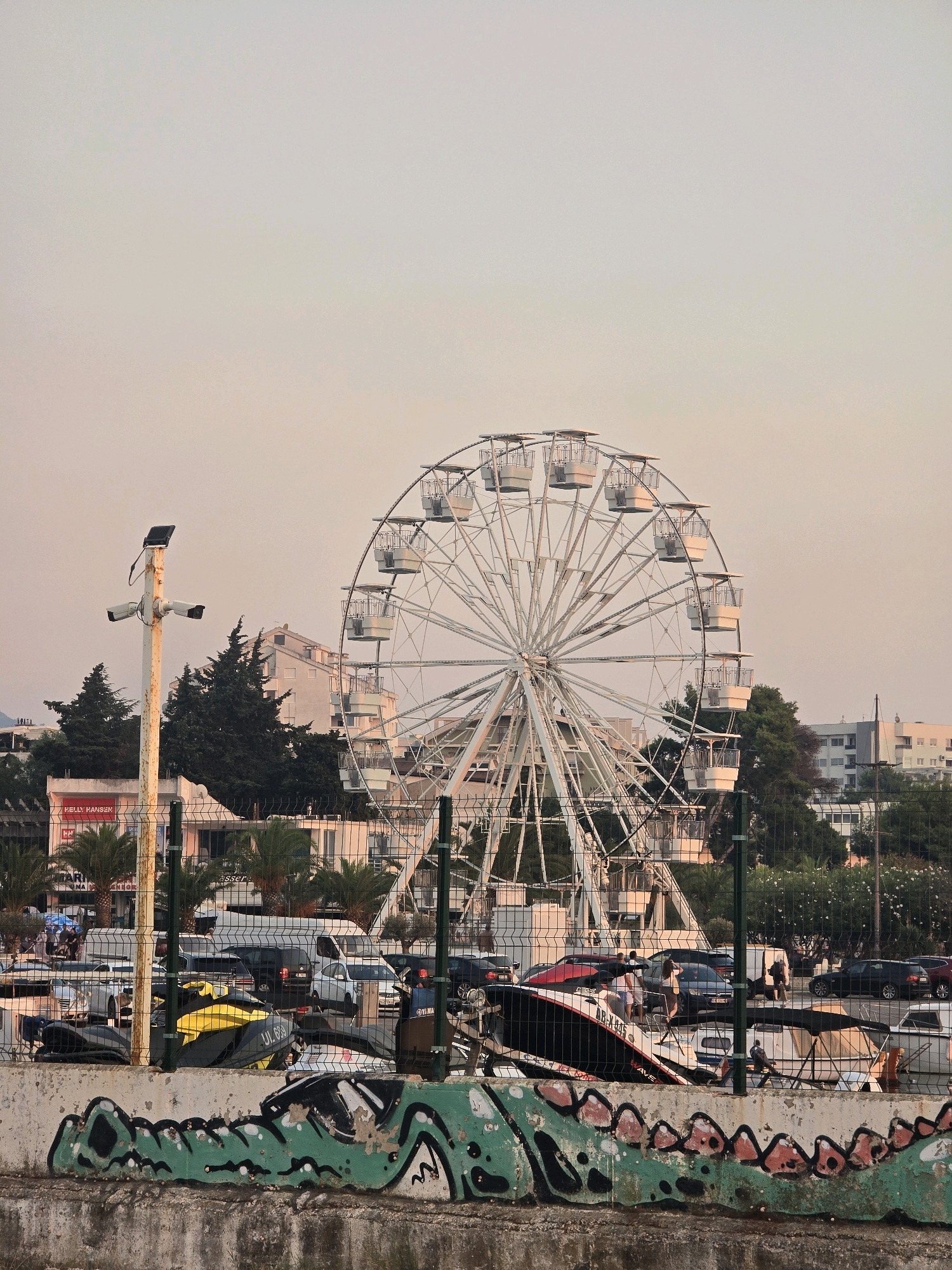 In the city of Bar, Montenegro, a large white Ferris wheel dominates the background of this scene, its intricate metal structure reaching towards the hazy, pastel sky. In the foreground, a concrete wall is adorned with graffiti depicting a green creature with red spikes. Behind this wall, a black metal fence encloses an area where various vehicles are parked, including cars and a yellow jet ski. A white pole with two security cameras is also visible. Further in the background, buildings with light-colored facades and dark-tiled roofs are nestled amongst lush green trees. The overall atmosphere is serene, with a hint of urban amusement.