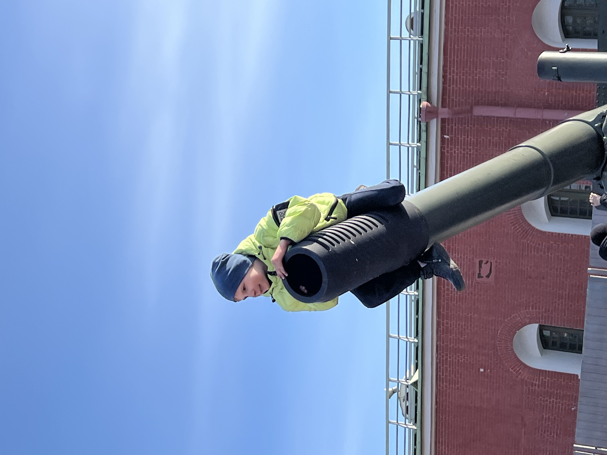A child, wearing a blue knit cap, a light green jacket, and dark pants and shoes, is sitting inside the barrel of a large cannon. The cannon is mounted outside a red brick building with arched windows and metal railings. The sky is bright blue. This is likely a historical site or museum. The location is the Peter and Paul Fortress in Saint Petersburg, Russia, known for its cannons and historic architecture.