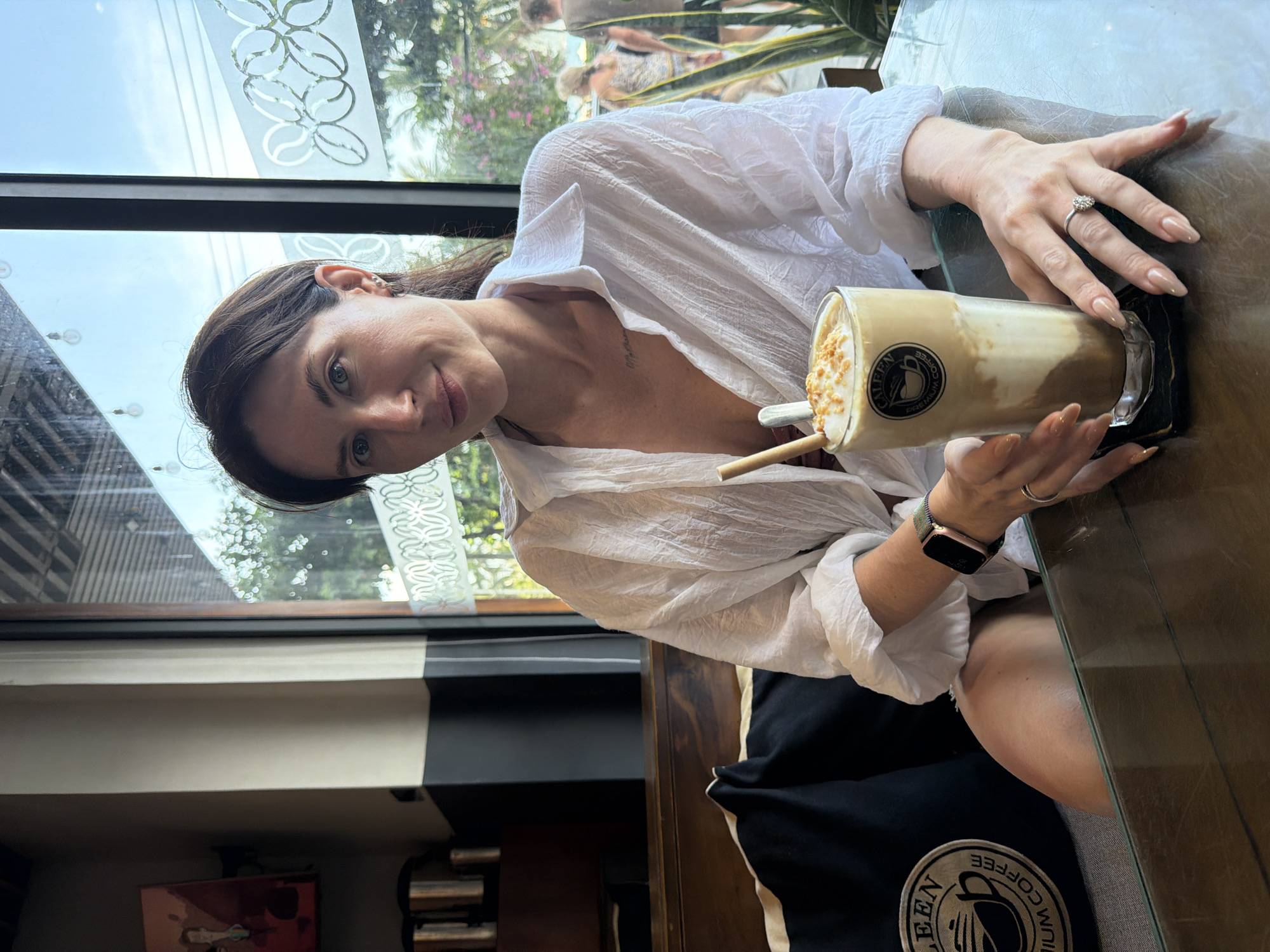 A woman is seated at a glass-topped table inside a Caleen Premium Coffee shop, likely located in Bac Nha Trang Ward, Vietnam, given the tropical outdoor setting. She is positioned towards the left of the frame, facing the camera with a subtle smile. She appears to be in her late 20s to early 40s, with dark brown hair and light-colored eyes. She wears a loose-fitting, textured white long-sleeved shirt, partially unbuttoned, revealing a glimpse of a darker garment underneath and a small tattoo on her upper chest. On her left wrist, she has an Apple Watch with a rose gold case and a dark mesh band. On the ring finger of her right hand, she wears a silver-colored ring with an oval-shaped clear gemstone. Her fingernails are long, almond-shaped, and painted a nude color.
In front of her, on a black coaster, is a tall glass containing a layered iced coffee drink. The drink features a dark brown bottom layer, a lighter brown/beige middle layer, and a white foamy top garnished with light brown crunchy bits. A bamboo-style straw and a small spoon are inserted into the drink. The glass displays the "CALEEN PREMIUM COFFEE" logo. Her hands rest on the table, with her right hand lightly grasping the glass.
The interior of the cafe features dark wooden accents, cream-colored walls, and dark grey paneling. To the left, a red-hued framed artwork hangs on the wall, and below it, several metallic containers are visible on a counter. The woman is seated on a bench with a dark cushion, behind which a black pillow with the "CALEEN PREMIUM COFFEE" logo is visible.
Behind the woman, a large window spans the width of the frame, allowing ample natural light and revealing an outdoor scene. The sky is clear and blue, and dense green foliage suggests a tropical environment. The window has a decorative frosted pattern along its mid-section. Beyond the window, indistinct figures of people are visible walking on what appears to be a street, along with more lush green plants immediately outside. Overhead, several bare light bulbs are suspended within the cafe.