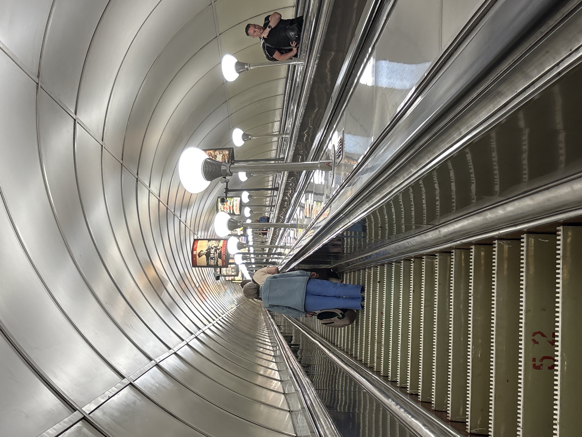 The image depicts an interior view of a very long escalator in the Saint Petersburg Metro system, likely within an art nouveau-designed station given the arched metallic ceiling. The escalator is flanked by polished metal railings and illuminated by a series of white, globe-shaped lights. On the escalator itself, several individuals are visible. In the foreground, a woman wearing a light blue denim jacket and blue pants is ascending, carrying a large bag. Further up, other passengers are also traveling upwards. On a parallel escalator to the right, a man in a black t-shirt and black shorts is standing, wearing a black bag over his shoulder. Posters are visible along the tunnel walls, advertising events or performances. The escalator steps are a pale green color with reflective metal edges, and some steps have red markings on them, such as the number "52". The overall scene is clean and well-lit, characteristic of public transportation infrastructure.