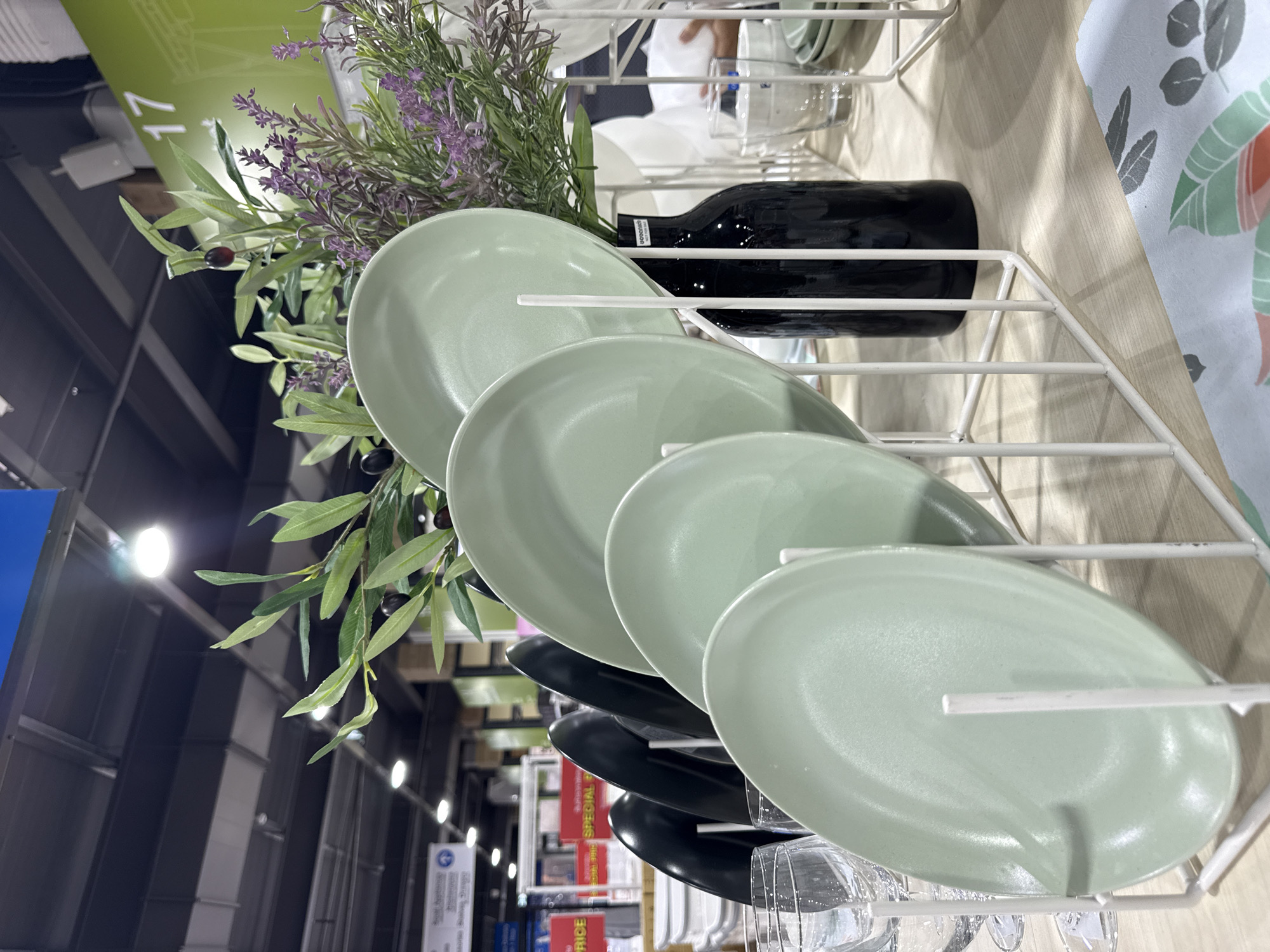 A close-up, eye-level shot captures a display of light green oval plates in a white metal rack at a retail store, possibly in Baan Chaweng Noi, Thailand. The plates are angled and stacked, with the largest at the front. Behind the plates, a dark, glossy vase holds artificial greenery with dark olives and sprigs of lavender. To the left, a few clear wine glasses are visible. In the background, blurred shelves with various items and a sign with red and white text suggesting a sale are discernible. Overhead, a dark ceiling with exposed beams and lighting fixtures is present, and a green sign with the number "17" is visible on the right. The lighting appears to be from artificial sources, creating some reflections and shadows. No people are clearly depicted in the immediate foreground or interacting with the displayed items.