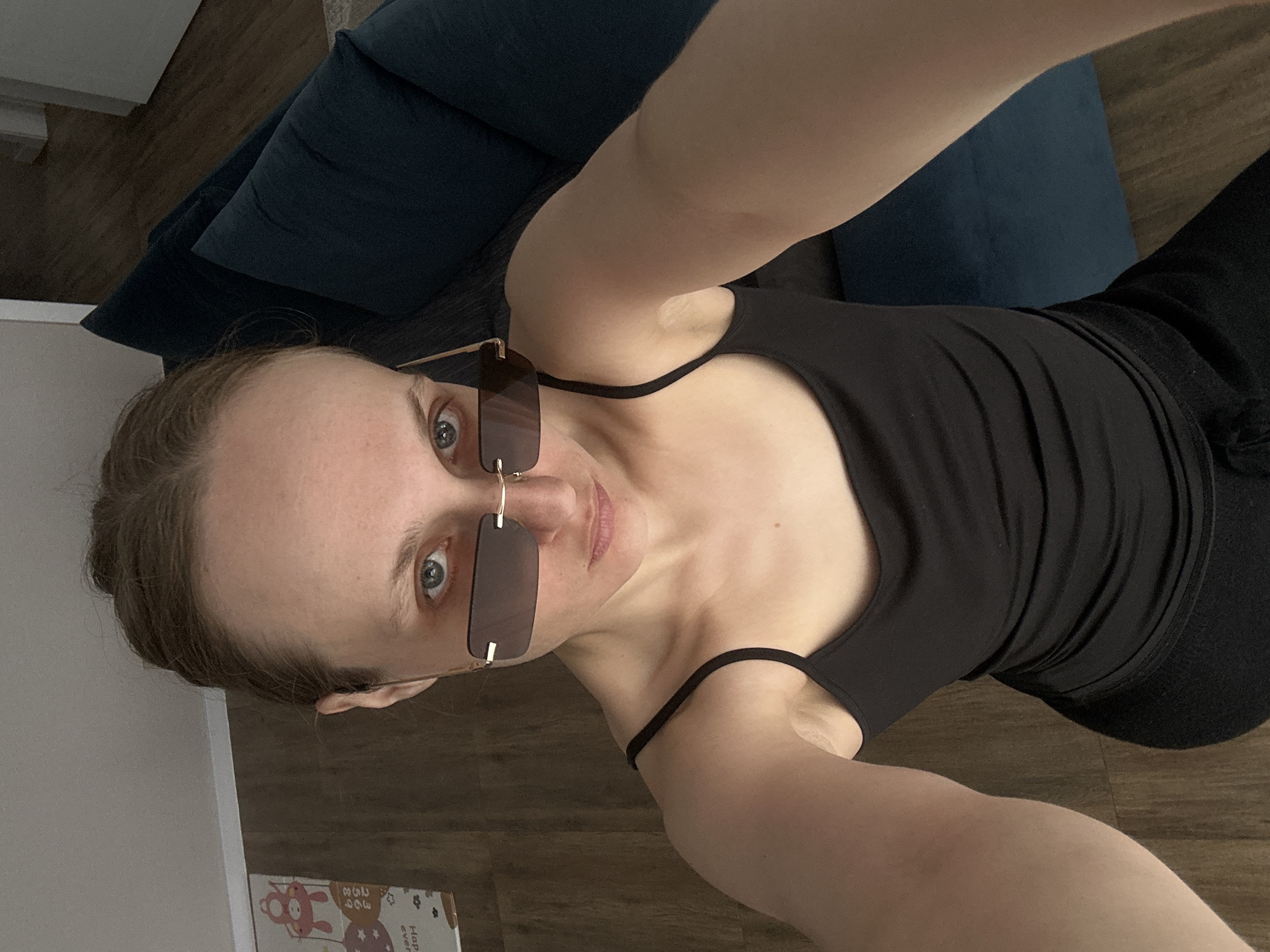 A selfie taken indoors from an elevated angle. A fair-skinned person with light brown hair pulled back is wearing sunglasses and a black tank top, with part of their black pants visible in the bottom right. They are positioned in front of a dark blue couch. To the left, there is a white cabinet and a partially visible rectangular object with cartoonish illustrations on a shelf. The floor appears to be light brown hardwood. The location is unknown and cannot be confirmed to be Sepych, Russia.