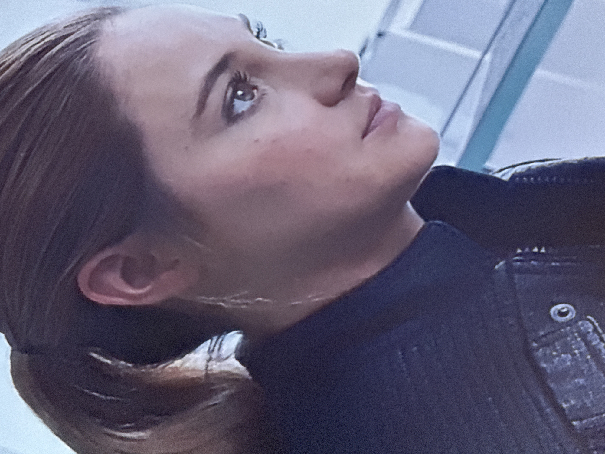 A close-up shot shows a woman with light brown hair pulled back into a ponytail. She is looking upwards and towards the left. Her features include light skin, a slightly prominent nose, and her left ear is visible. She is wearing a dark-colored collared jacket with a visible button detail. The background appears to be a light-colored or white interior space with linear elements suggesting architectural features. There is no verifiable information to place the scene within Mytishchi, Russia.