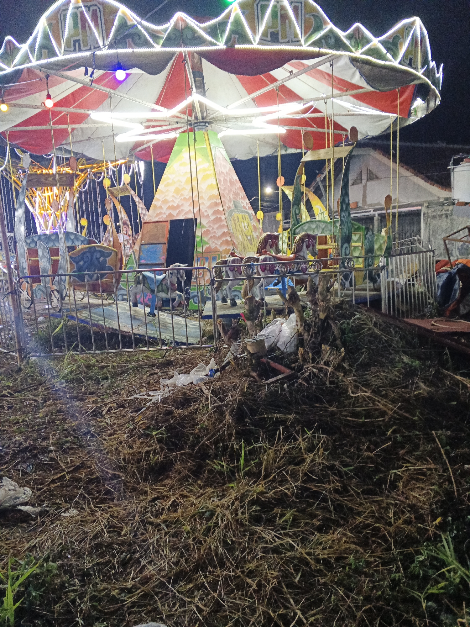 The image captures a carousel ride illuminated by decorative lights at night. The carousel features a conical structure at its center, painted with a mountainous landscape. Various animal figures, including horses and giraffes, are visible as part of the ride's attractions. A metal fence surrounds the carousel, separating it from the foreground, which is covered with dry grass, branches, and some scattered white plastic debris. The overall scene suggests a temporary amusement setup, possibly at a local fair or event, with a slightly neglected or unkempt surrounding area. The location is identified as Sukabumi, Indonesia. There are no people visible in the image.