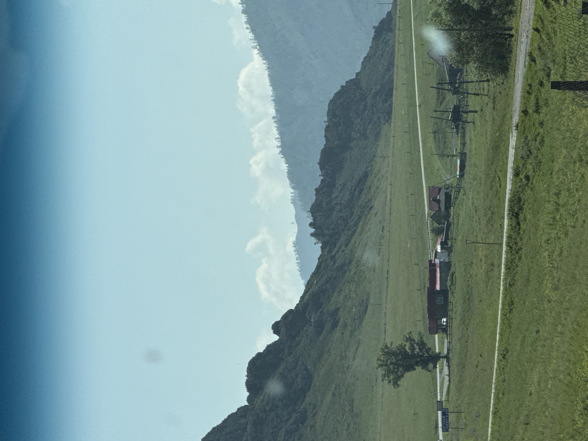 The image depicts a rural landscape with rolling green hills and mountains in the background. A road winds through the scene, passing by several buildings with red roofs. A lone tree stands in a grassy field, and a fence can be seen in the foreground. The sky is clear and blue. Based on the sign which reads "Туюкта 18 Ташанта 323" (Tyukta 18 Tashanta 323), the location is likely near the village of Tyukta, Russia, on the Chuya Highway. The sign indicates distances to Tyukta and Tashanta. The scene appears to be tranquil and undeveloped, typical of a remote area. There are no people visible in the image.