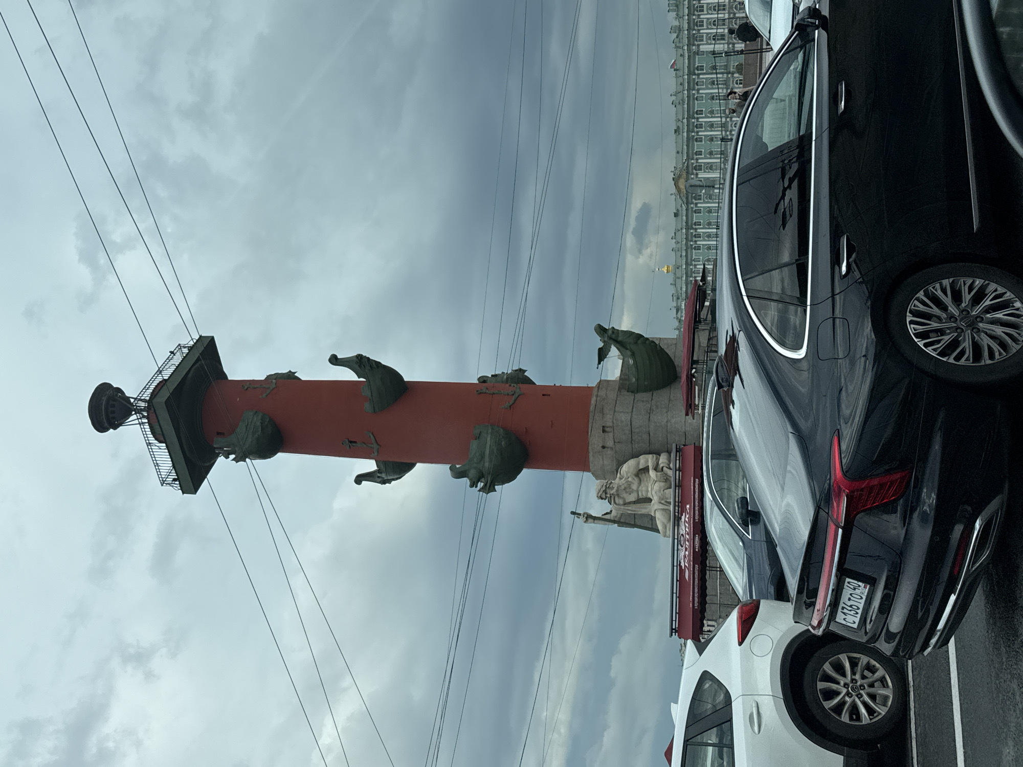 The image captures the Rostral Column on Vasilievsky Island in Saint Petersburg, Russia. The sky is overcast with dark clouds. In the foreground, several cars are parked, including a white SUV on the left and a black sedan in the center. The black sedan displays a Russian license plate that reads "C 136 TO 40". Behind the cars, the Rostral Column, a prominent landmark, stands tall. It is a red cylindrical structure adorned with sculptures of ships and anchors, and topped with a lantern. At the base of the column, a large stone sculpture of a seated male figure, possibly Neptune, holding a trident, is visible. In the background, a long, green classical building with a golden dome can be seen, which is likely the General Staff Building or part of the Hermitage Museum complex. A few people are walking in the distance. The overall scene suggests an urban environment with historical architecture and modern traffic.